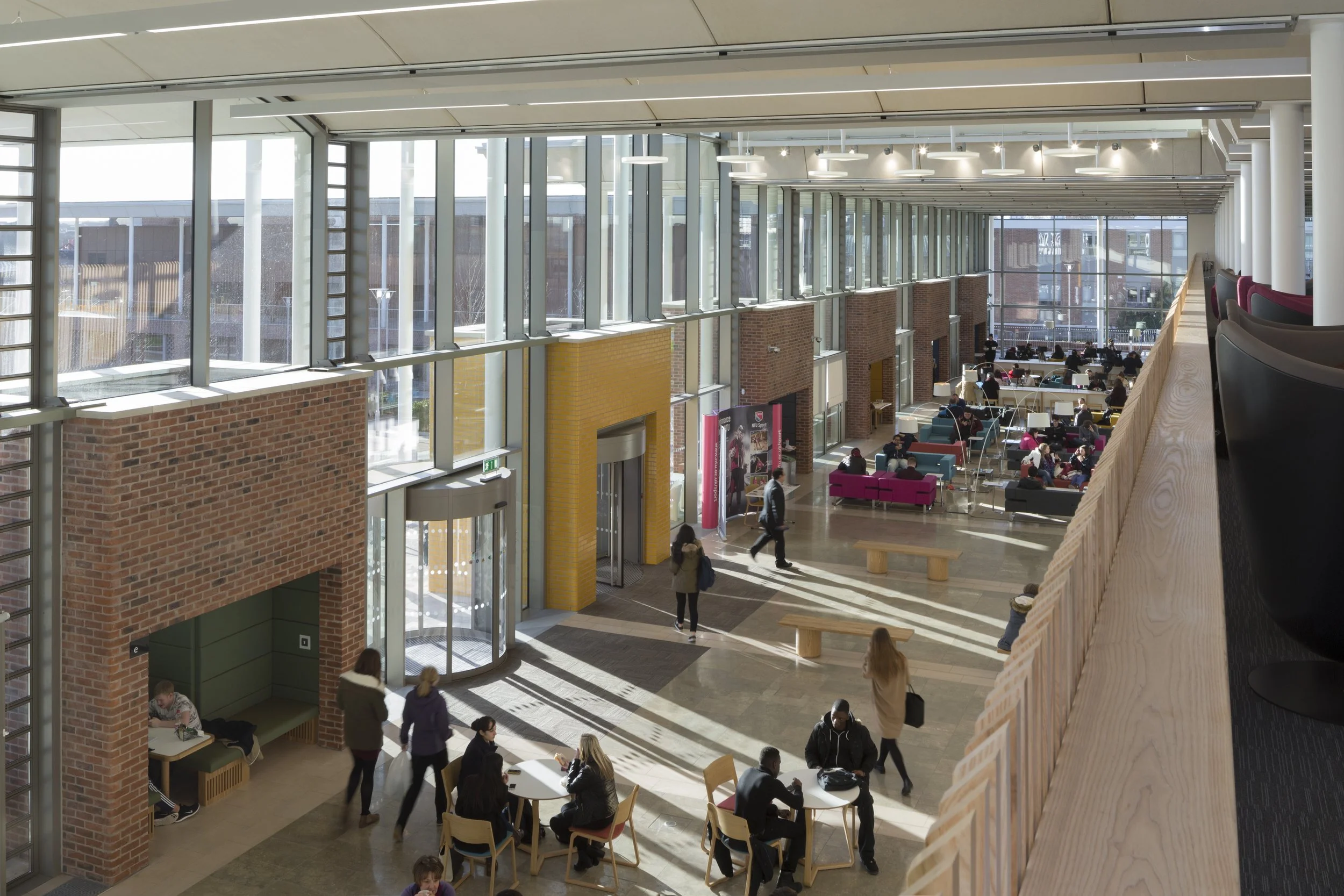 The interior of a modern, spacious building lobby or common area with large windows, brick walls, multiple seating areas, and people walking, sitting, and working.