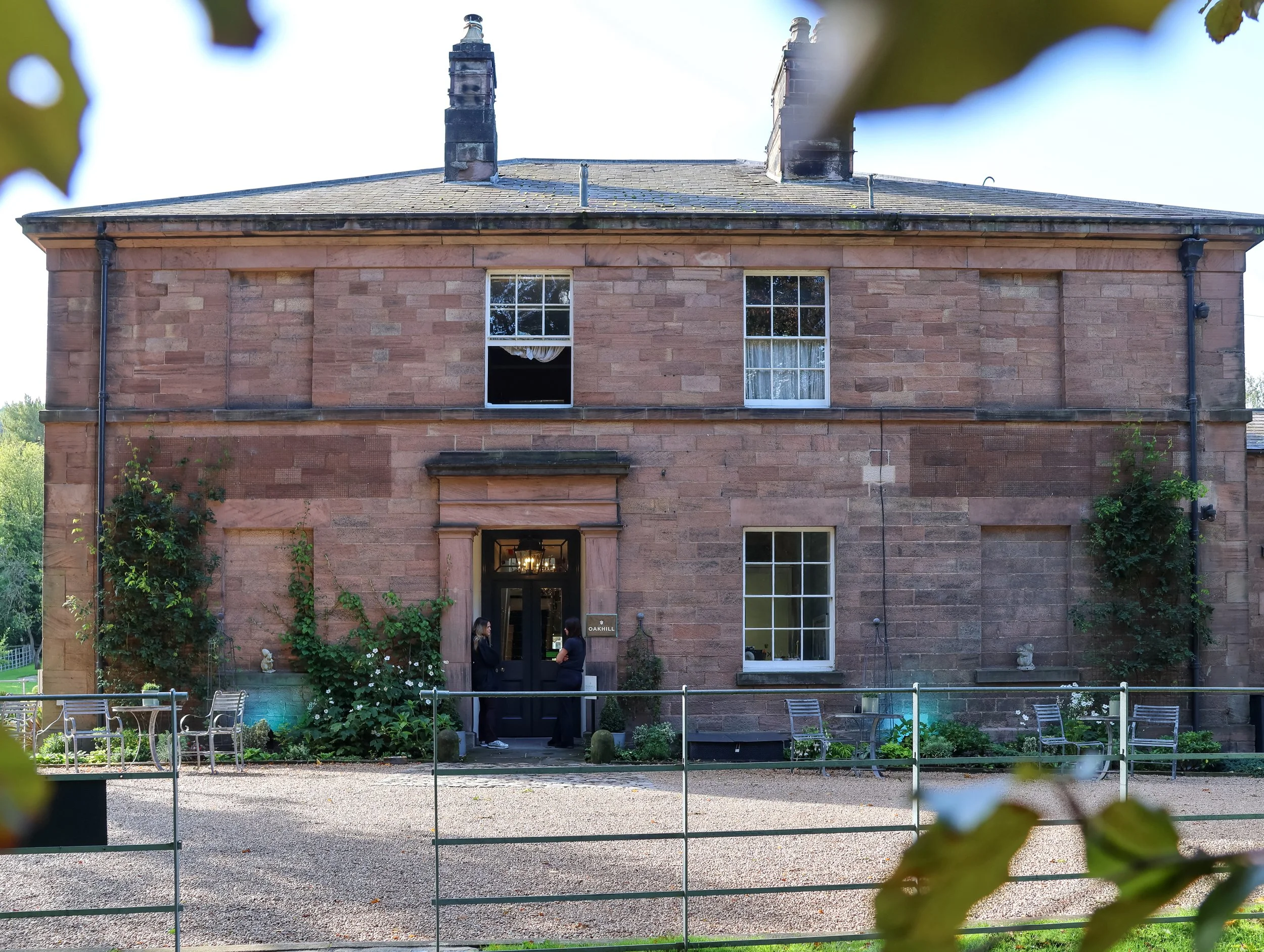 A brick building with multiple windows and a door, surrounded by a garden with greenery and bushes, and a metal fence in the foreground. Two women are standing near the entrance, engaging in conversation, with chairs and tables visible outside.