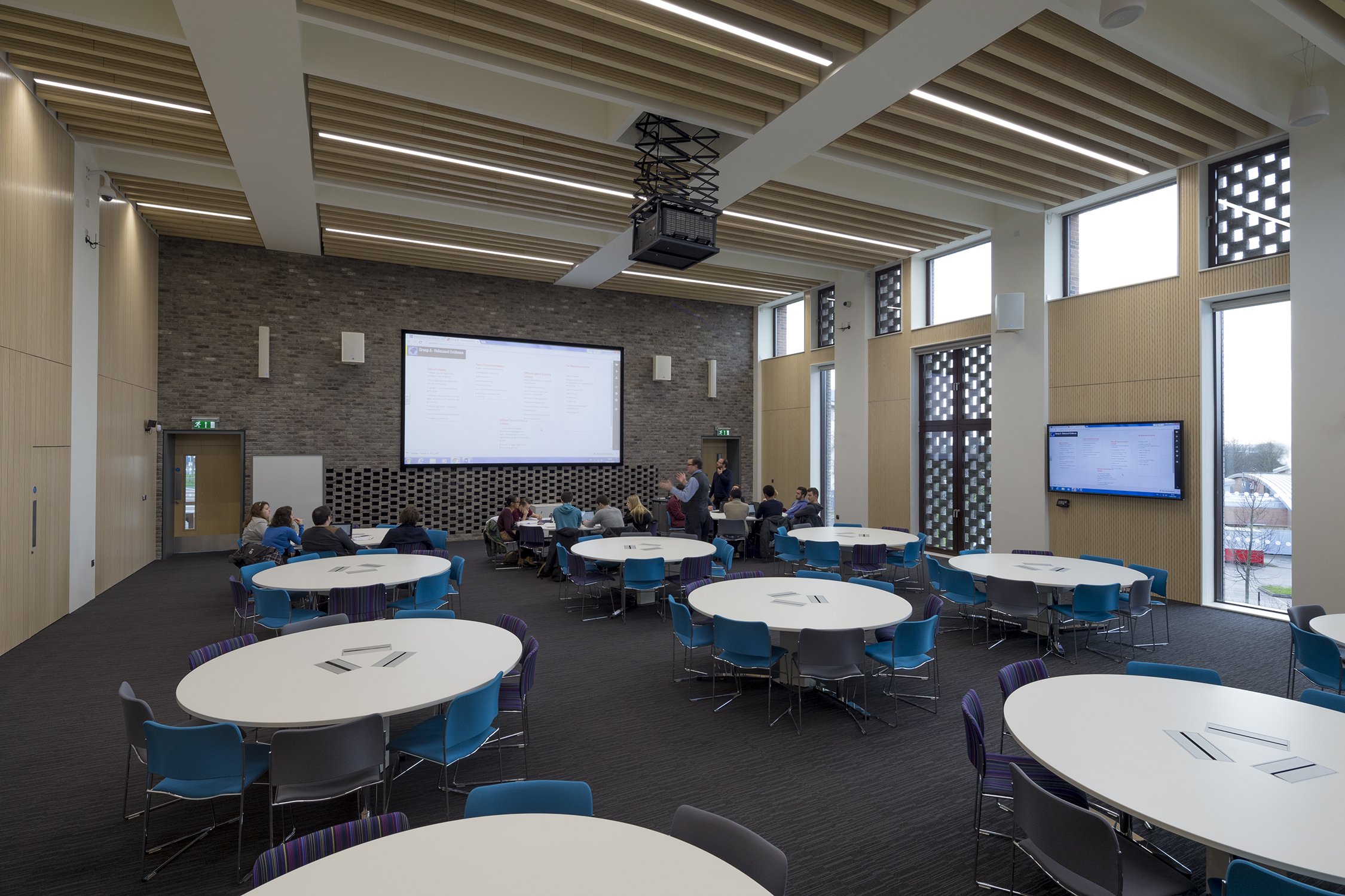A modern conference room with round tables and colorful chairs, large windows, a brick accent wall with screens, and wood ceiling panels.