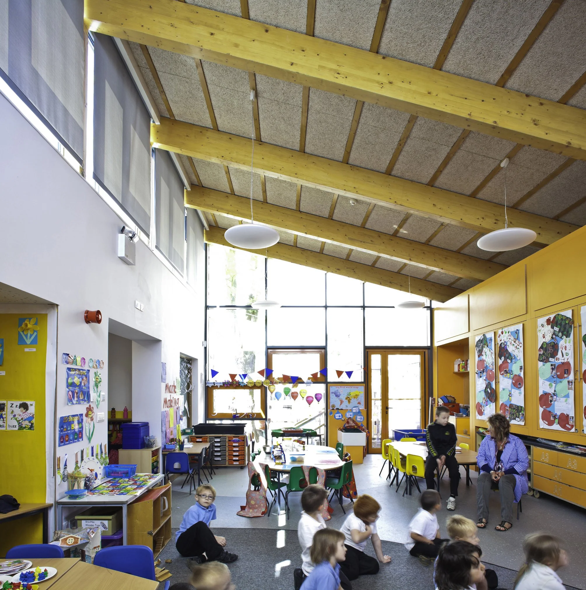 A classroom with children sitting on the floor and desks, decorated with colorful posters, art, and bunting. Large windows let in natural light, and there are wooden beams on the ceiling.