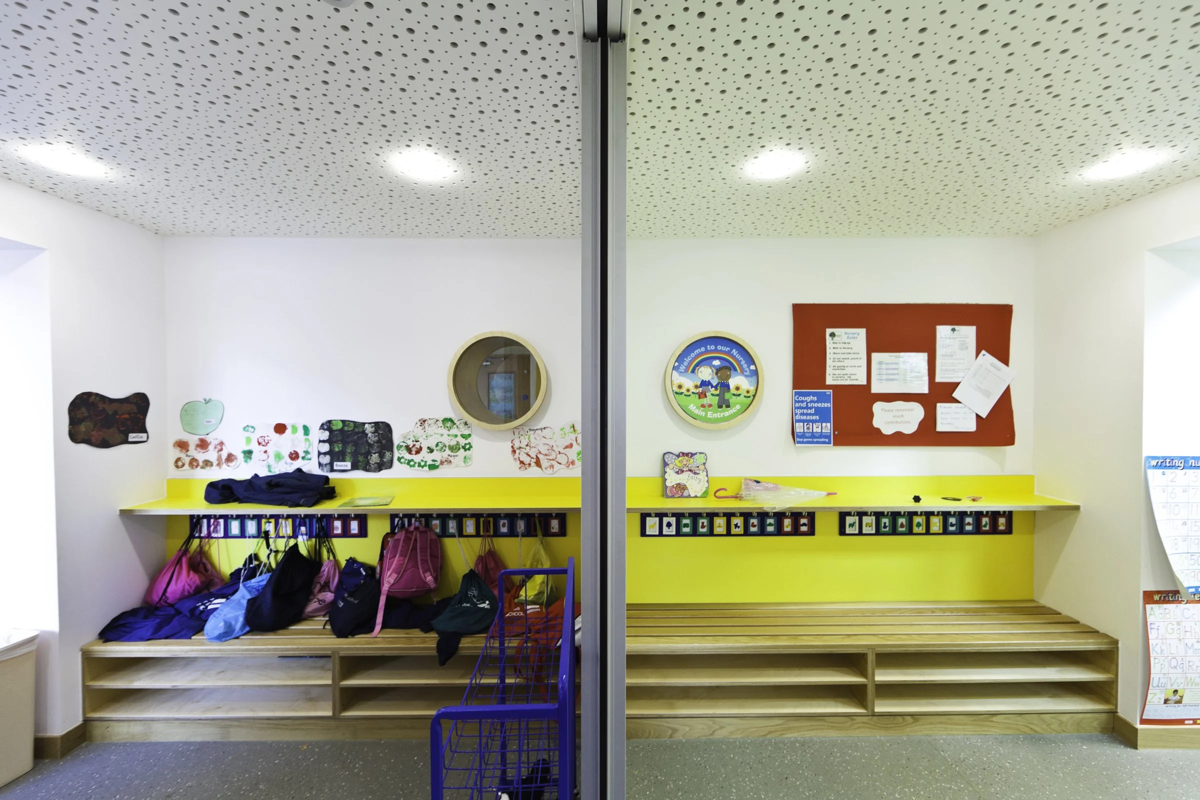 A children's classroom with backpacks hanging on hooks and colorful artwork on the walls. The room has a split mirror door reflecting the opposite side of the classroom, which contains a red bulletin board with notices and a yellow shelf with childre