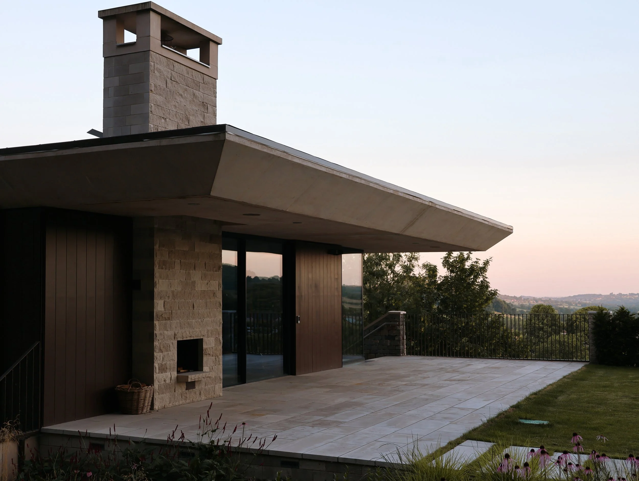 A modern house with a combination of stone, wood, and concrete exterior, featuring a large chimney and a spacious patio with tiled flooring, surrounded by a railing and overlooking a scenic landscape at dusk.