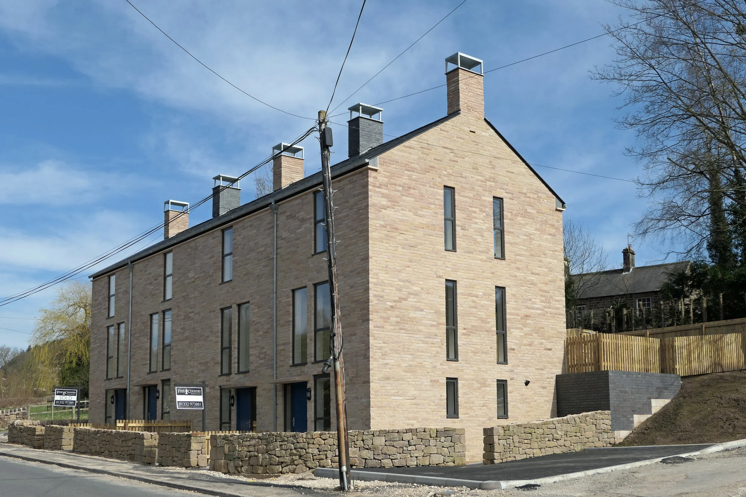 A modern four-story brick building with large vertical windows. There are signs indicating units for sale or rent on the ground floor. The building is located on a curved road with a stone wall and wooden fencing around the property. Utility wires an