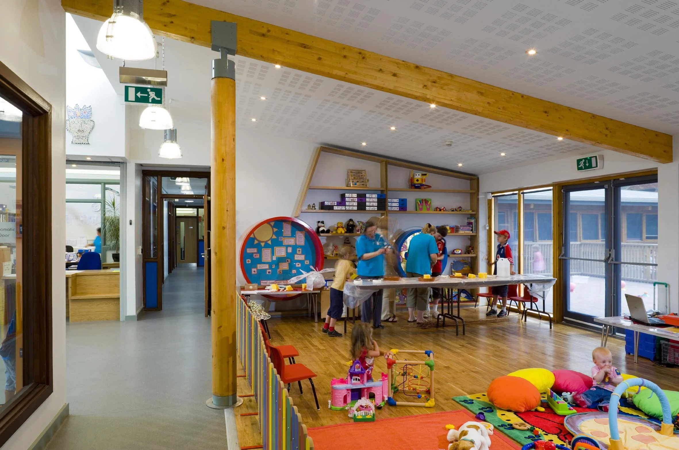 Children playing in a brightly lit indoor playroom with shelves of toys, a colorful rug, and large windows.