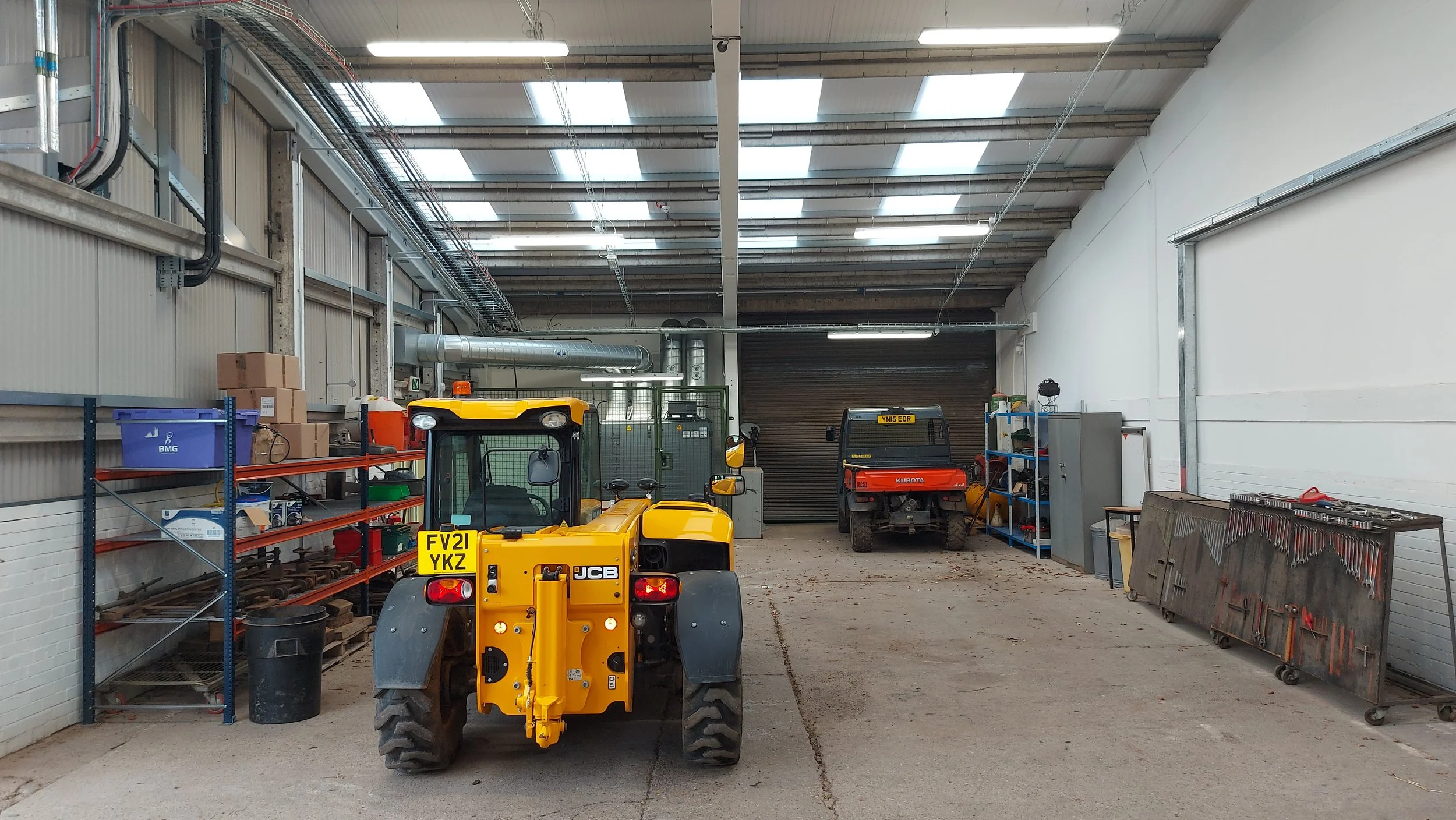 Inside a warehouse or garage with a yellow forklift and a red utility vehicle. Shelves with boxes and tools on the sides, industrial lighting, and a roller door in the background.