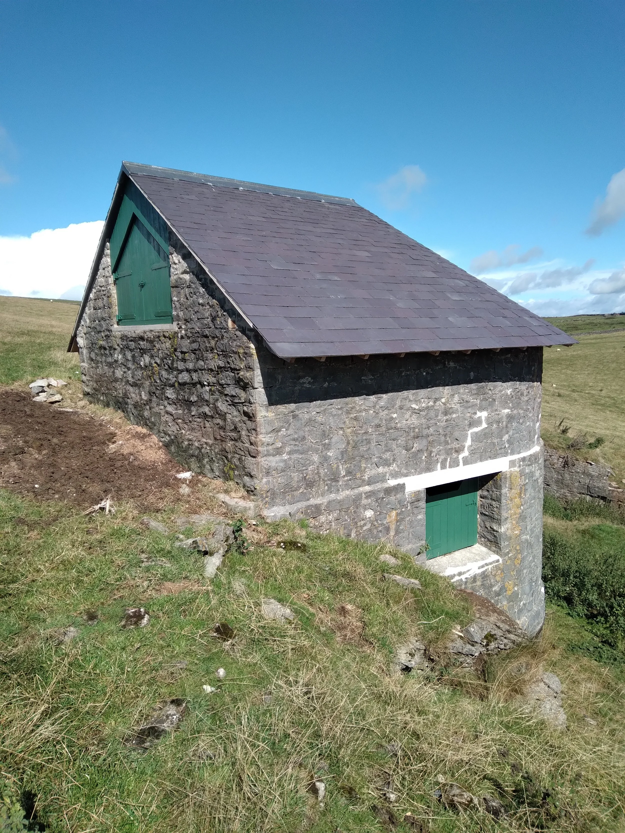 A small stone building with a steep roof and green shutters on a rural hillside under a blue sky with scattered clouds.