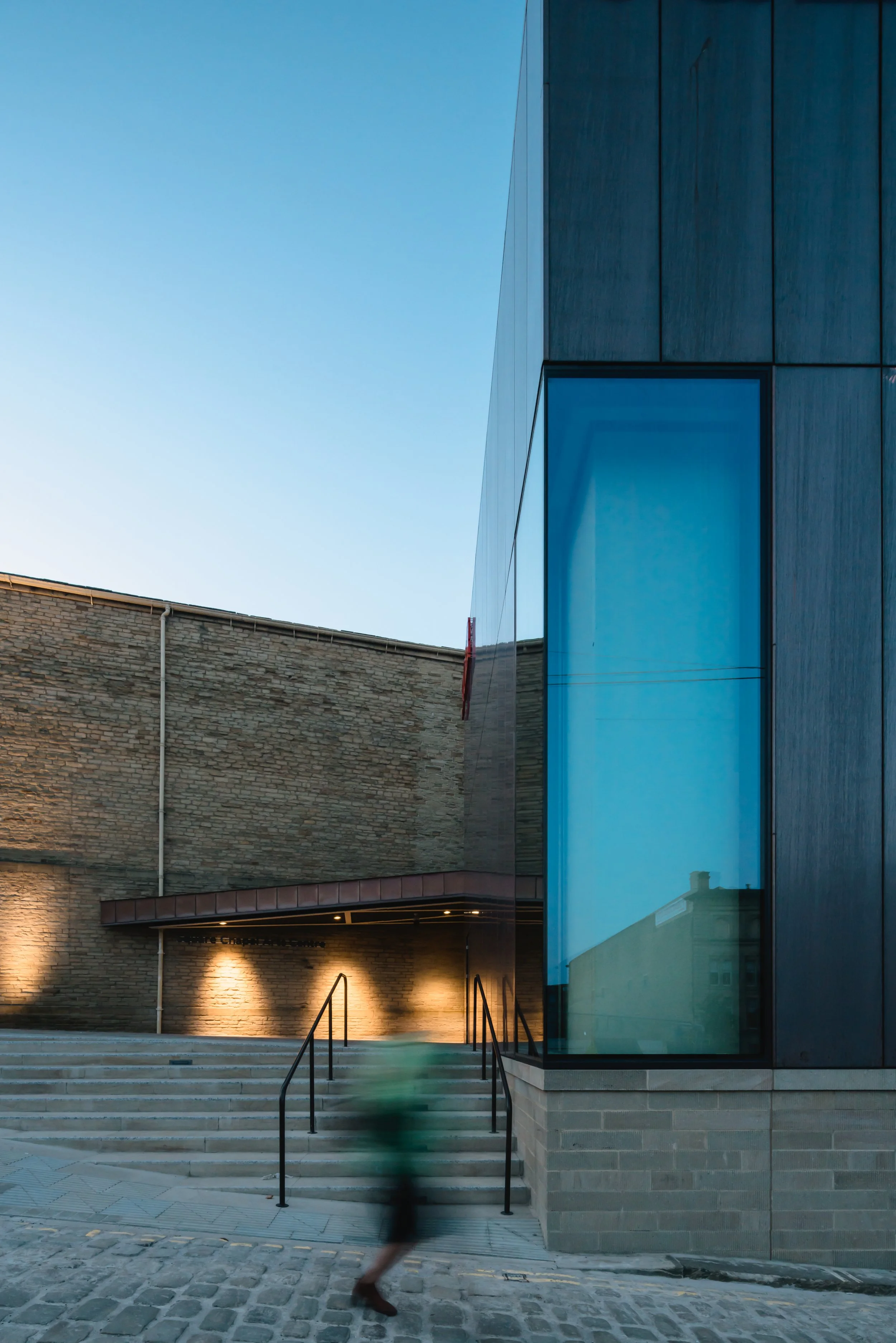 Modern building with large glass window, stone wall exterior, illuminated steps, and a person walking down the stairs.