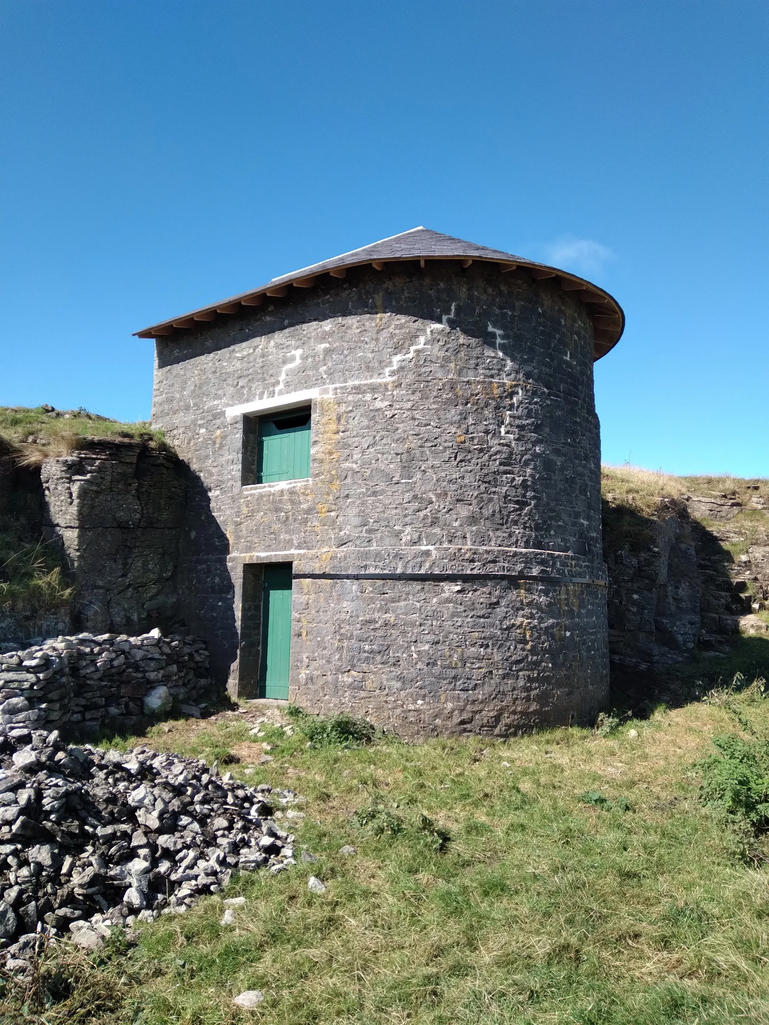 A stone tower with a green door and a small window, surrounded by grass and rocks, against a bright blue sky.