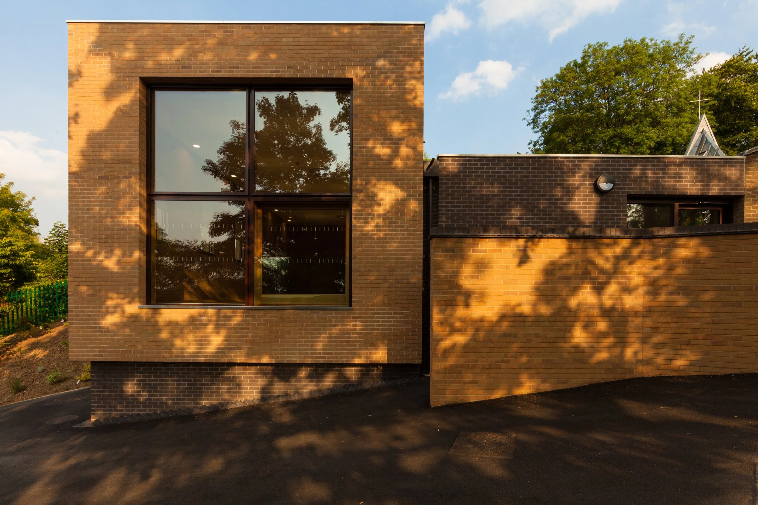 Modern brick building with large glass windows reflecting trees, situated outdoors during sunset with shadows on the facade and a clear blue sky.