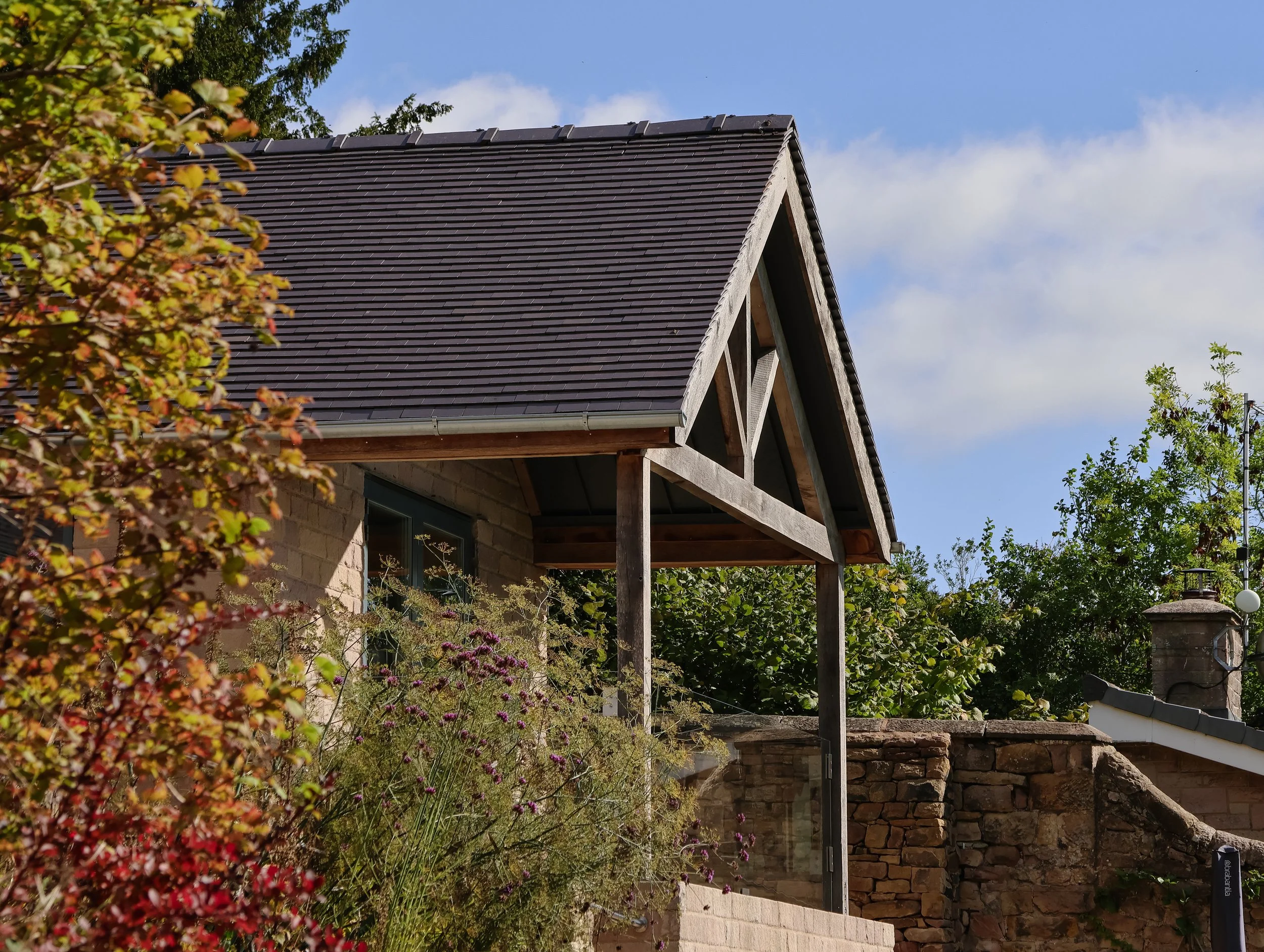 The image shows part of a house with a dark brown sloped roof, a brick wall, and a partially constructed or renovated exterior. There are trees and plants surrounding the house, with a blue sky and some clouds above.