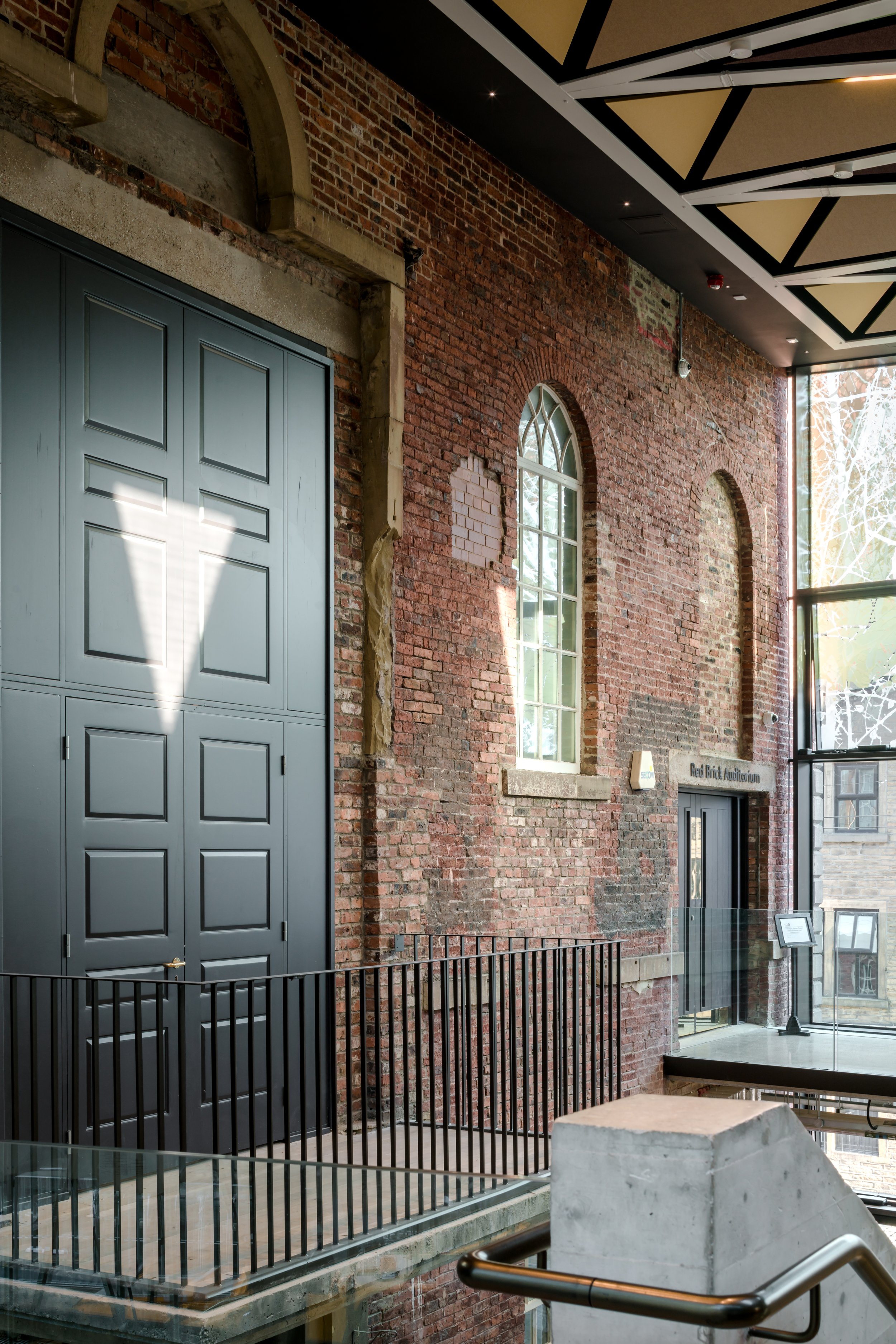 Interior view of a brick building showcasing tall arched windows, a black metal railing, and a modern ceiling with geometric patterns, with natural light coming through the windows.