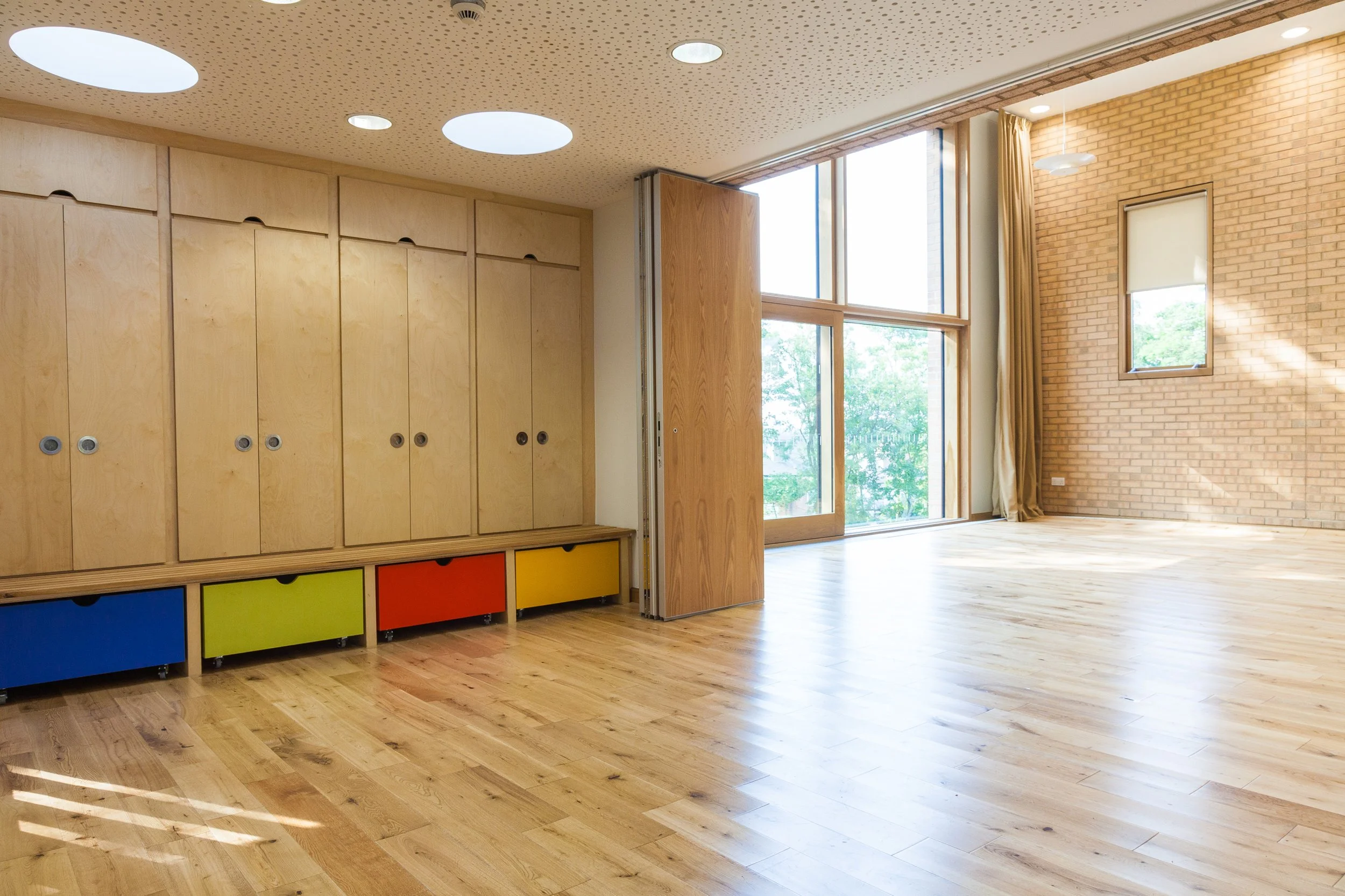 A spacious room with large windows letting in natural light. There are light wooden cabinets with colorful drawers underneath and a wooden partition wall. The ceiling has round skylights and small recessed lights. The floor is made of polished wood, 