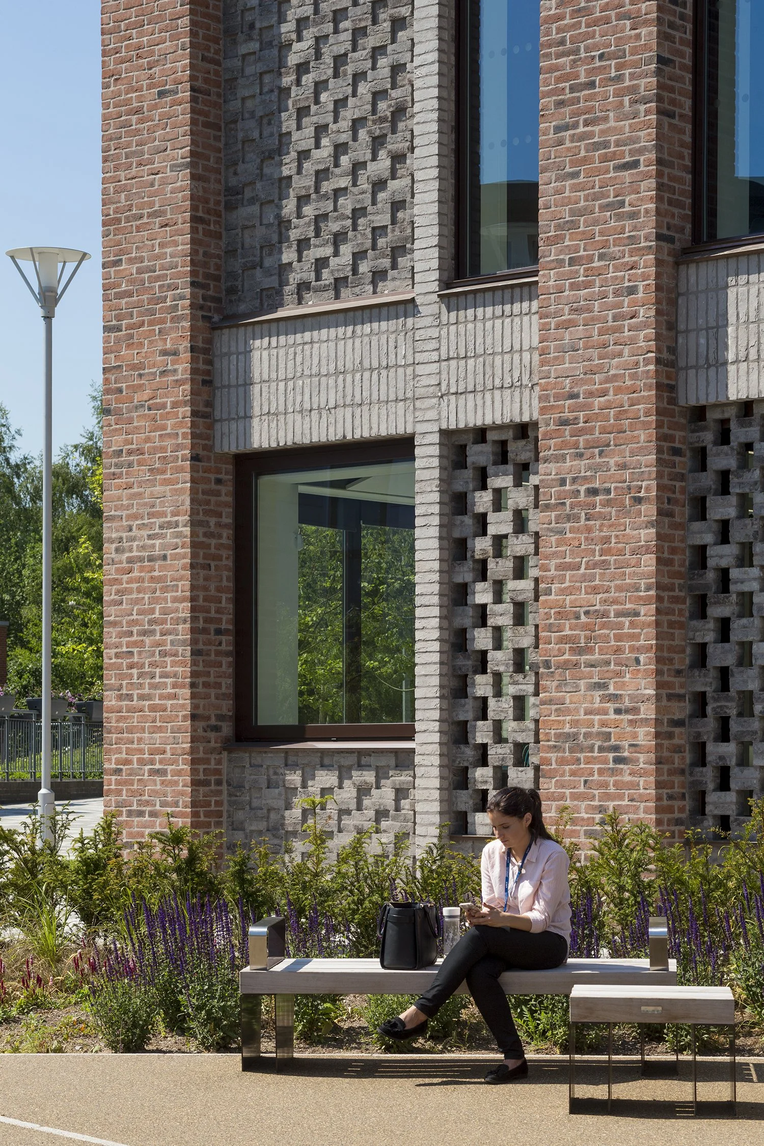 A woman sitting on a park bench looking at her phone, with plants and flowers in front, and a modern brick building with large windows behind her.