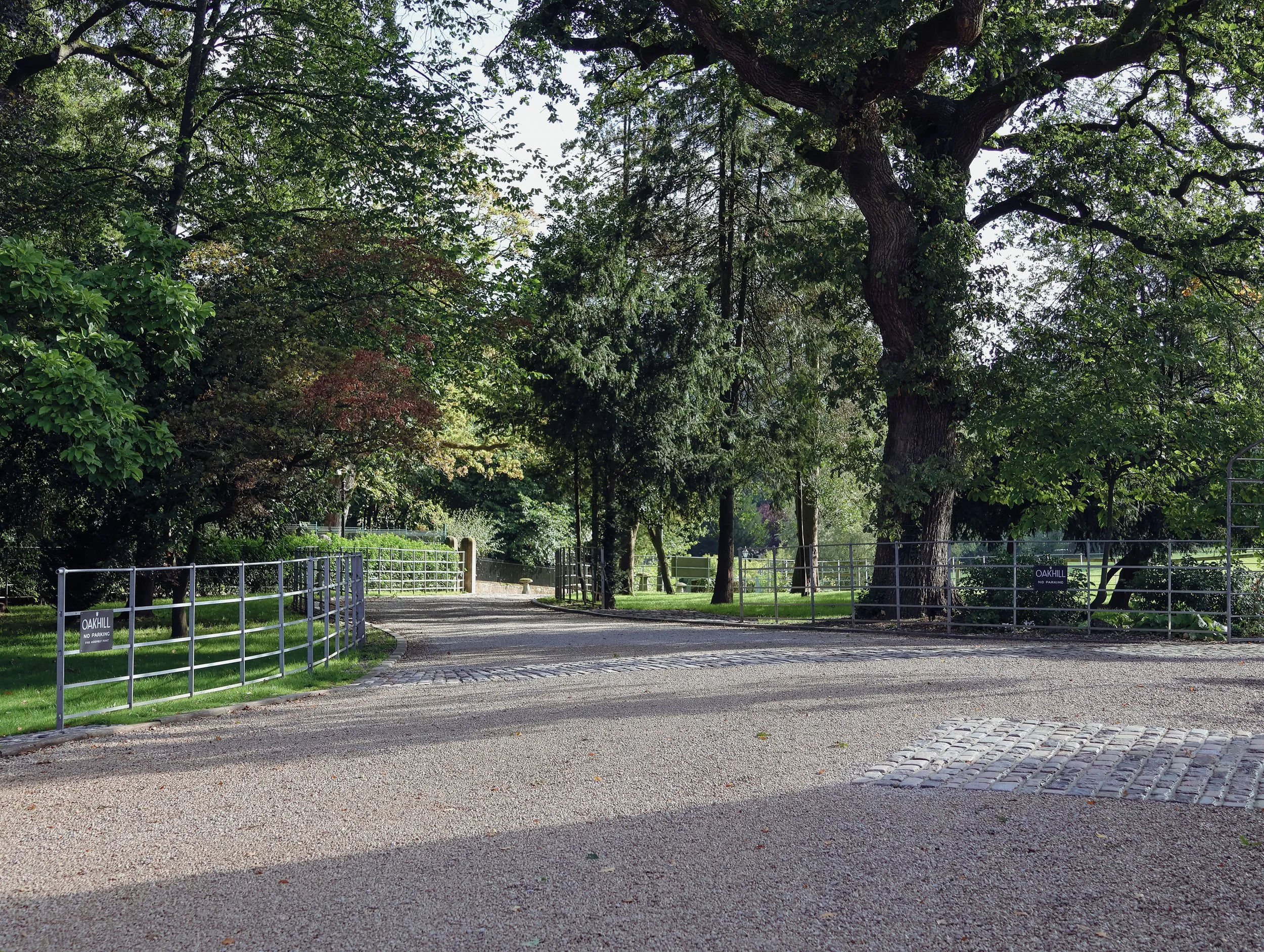 Park pathway with metal fence and green trees, sign reading 'Oakhill No Parking' in the background.