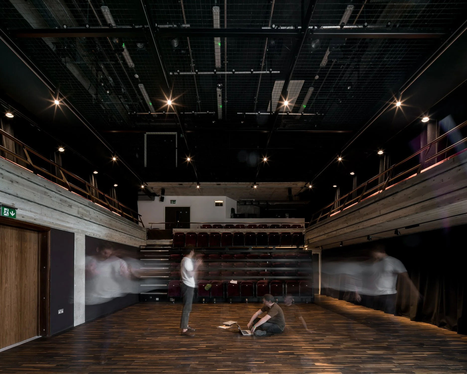 An empty theater or auditorium with a wooden floor, dark walls, a balcony with red seats, and a ceiling with lighting fixtures. There are a few blurred figures moving inside, some sitting on the floor near laptops.
