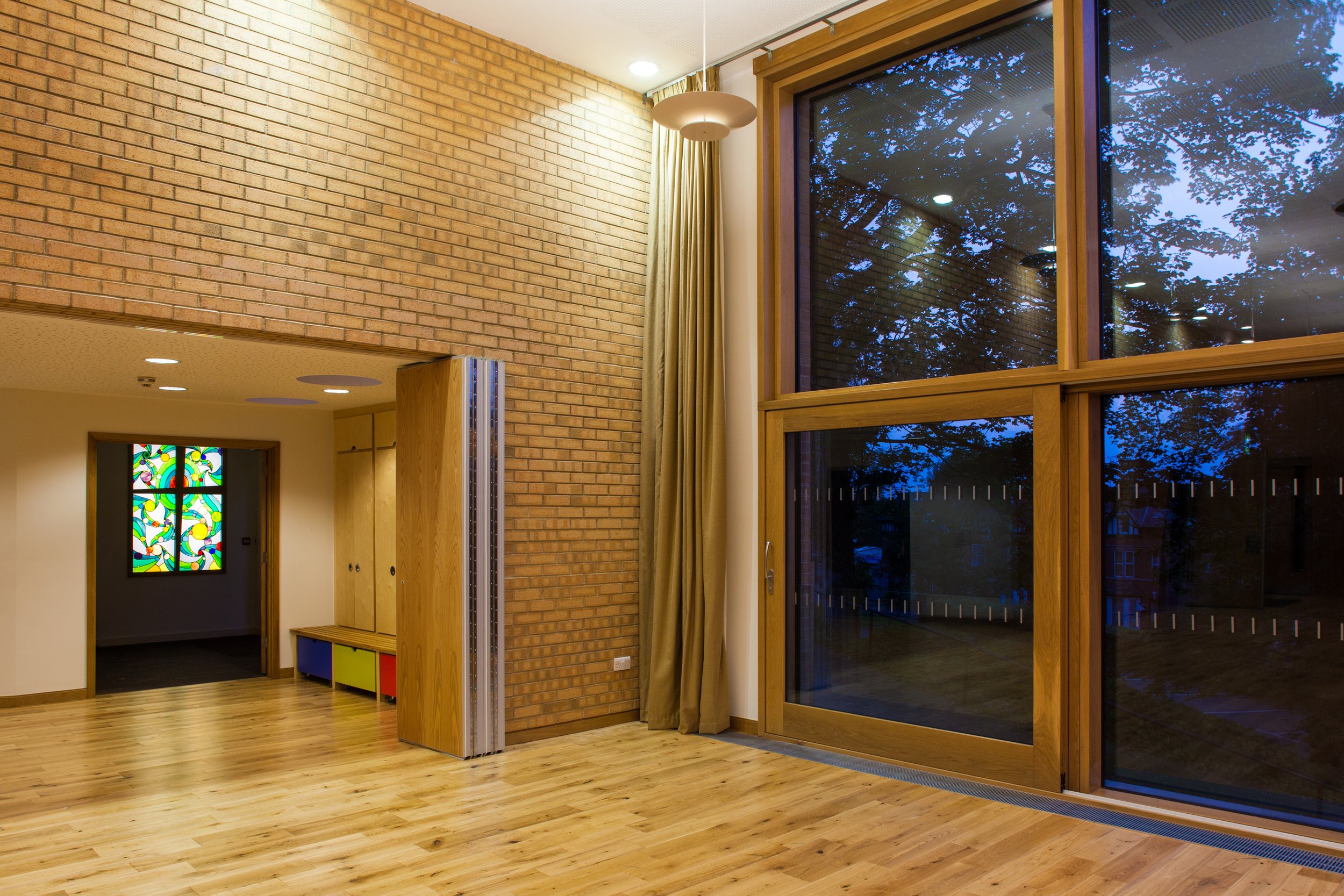 An interior room with wooden floors, brick wall, large windows with wooden frames, and beige curtains. There is a ceiling light and a colorful stained glass window in the background.