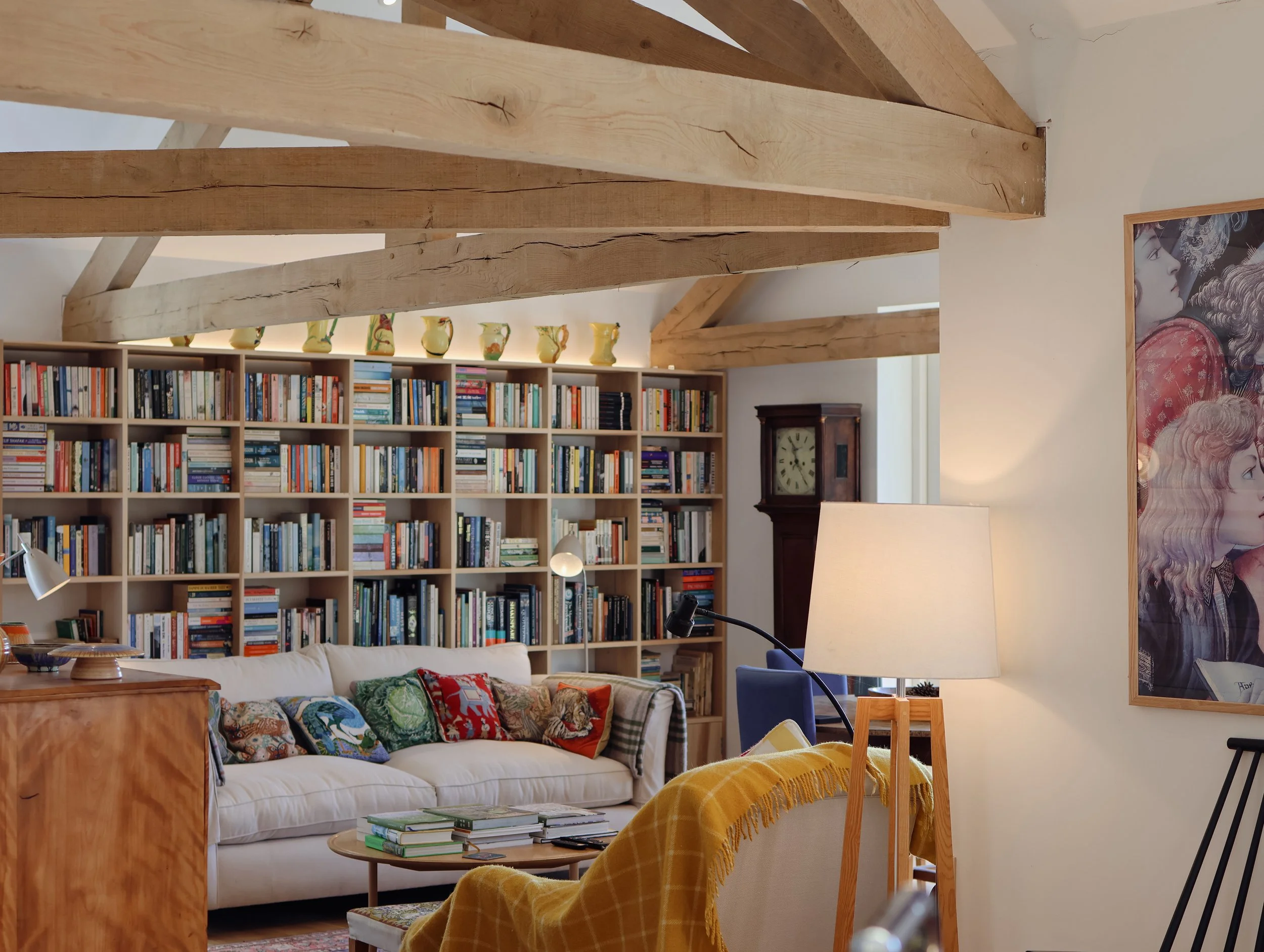 A cozy living room with a white sofa decorated with colorful pillows, a wooden coffee table with magazines, a tall bookshelf filled with books, a large portrait artwork on the wall, and exposed wooden beams on the ceiling.