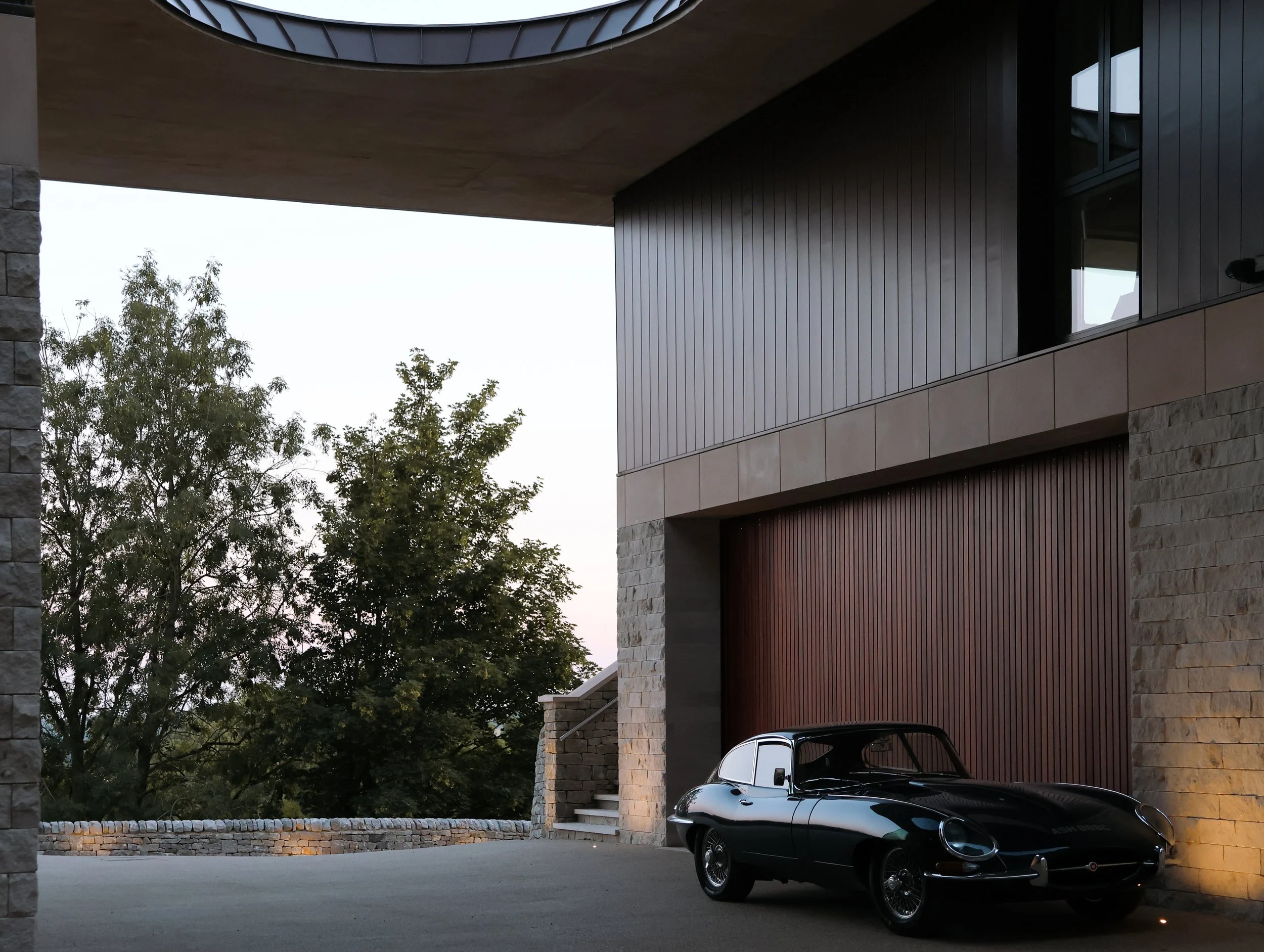 A black sports car parked on a driveway in front of a modern house with stone and wooden exterior features. Trees are visible in the background, and the sky is overcast.