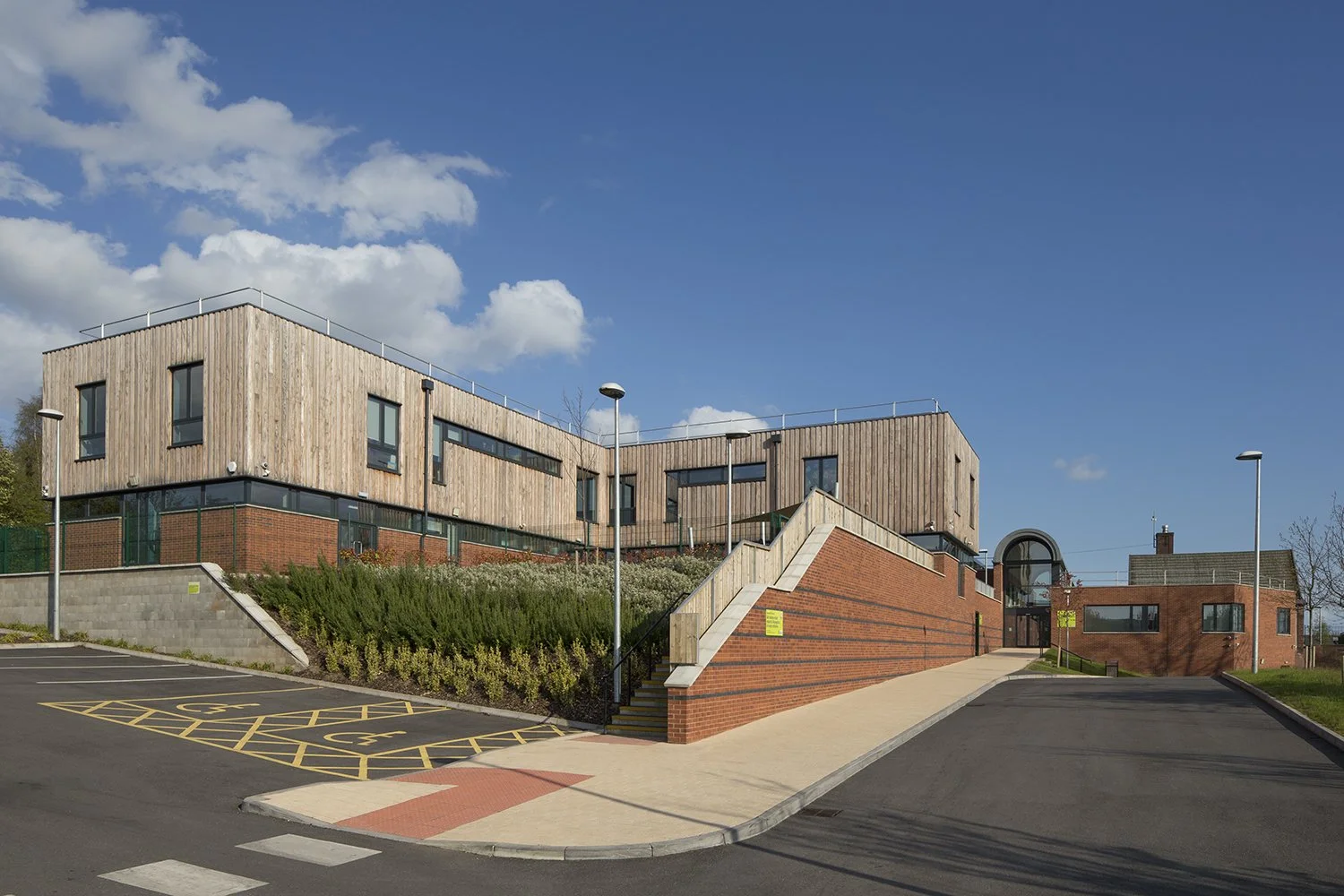 Modern building with wooden and brick exterior, accessible parking spaces, stairs, and pathways under a partly cloudy blue sky.