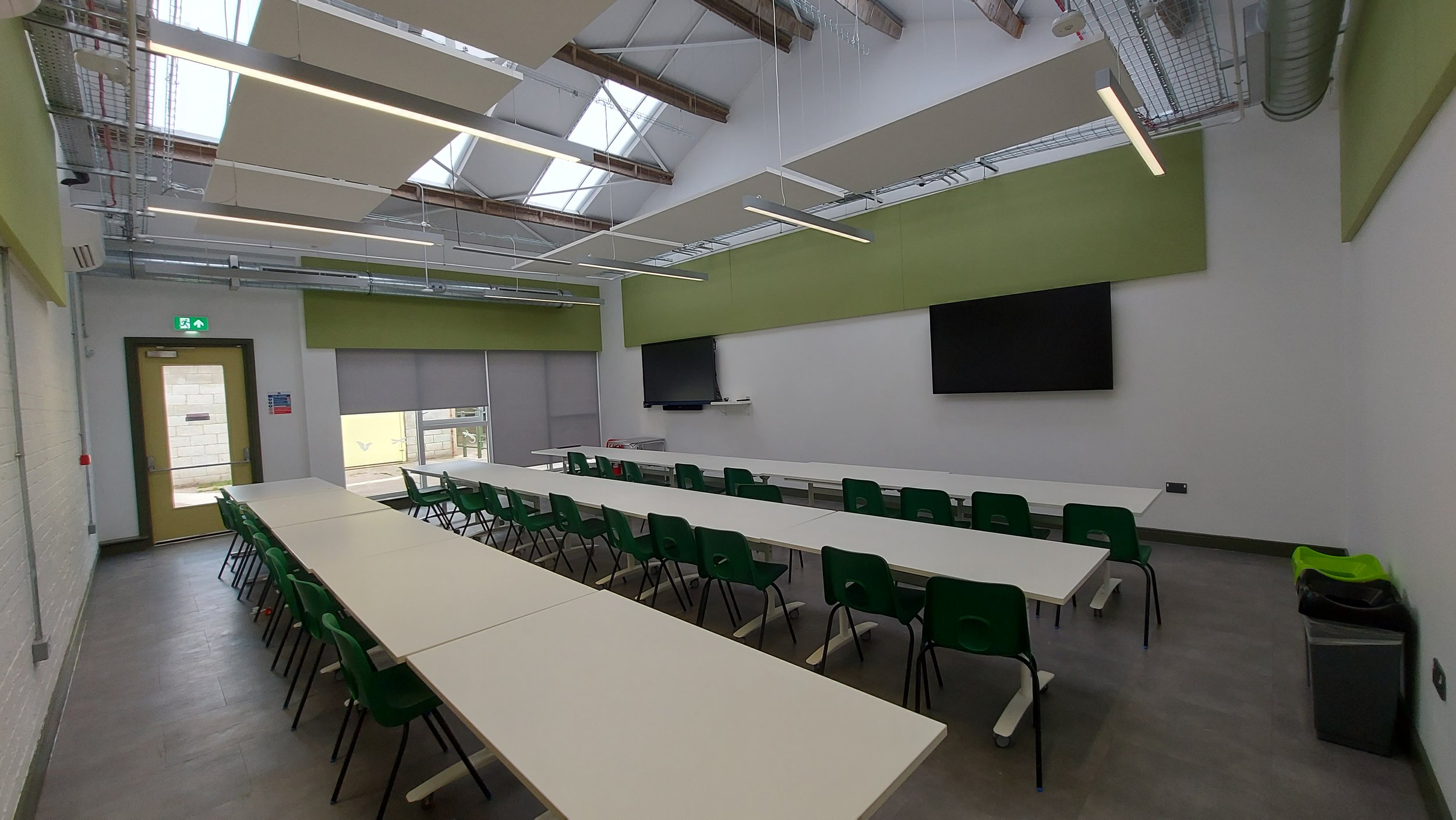 A classroom or meeting room with white tables arranged in a U-shape and green chairs. The room has a high ceiling with skylights, exposed beams, and white acoustic panels. There are two large flat-screen TVs on the wall and a door with a green exit s