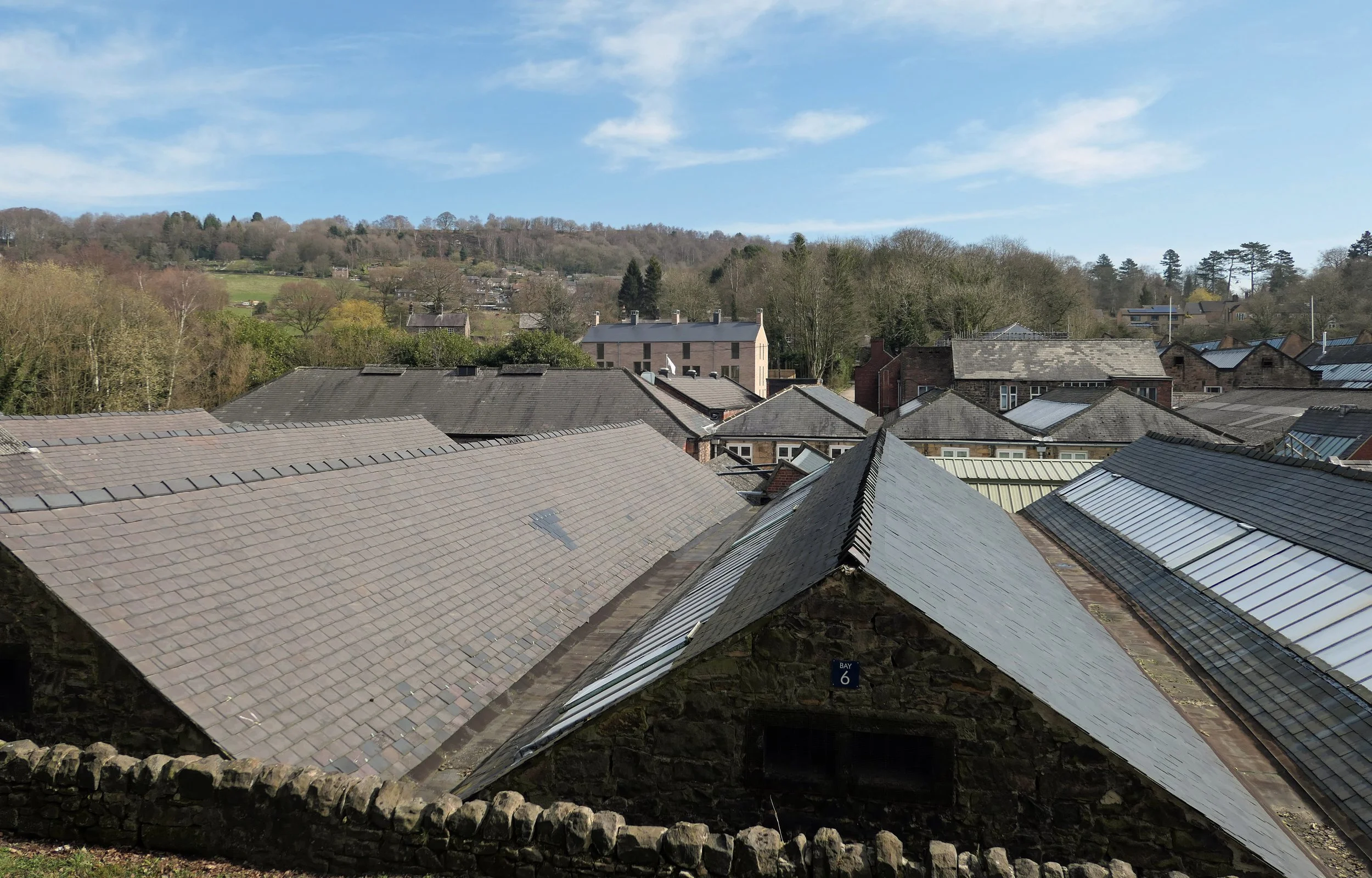 View of rooftops with slate tiles in a small town, surrounded by trees and hills under a blue sky with a few clouds.