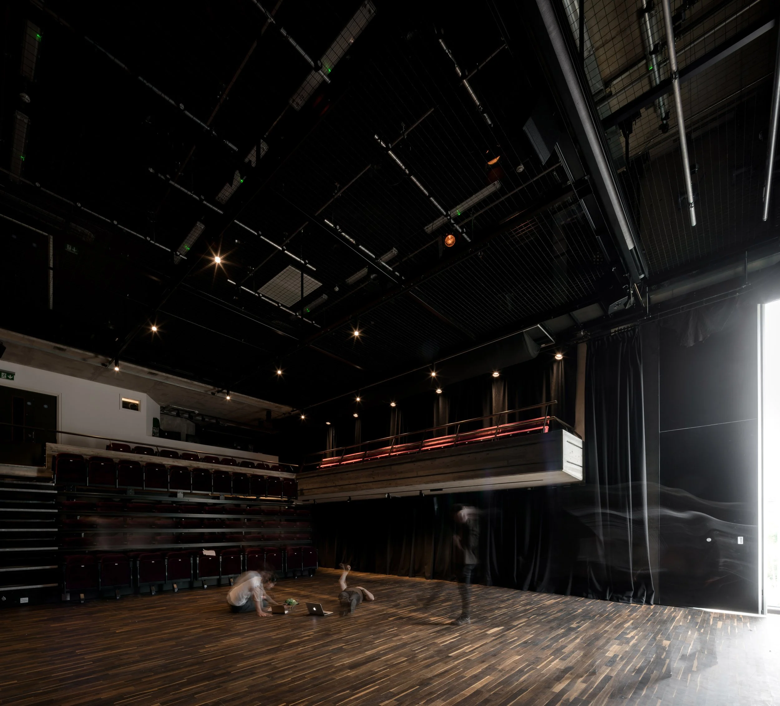 Empty theater or auditorium with rows of red seats, a wooden floor, a black ceiling with stage lights, and a few children sitting on the wooden floor near laptops.
