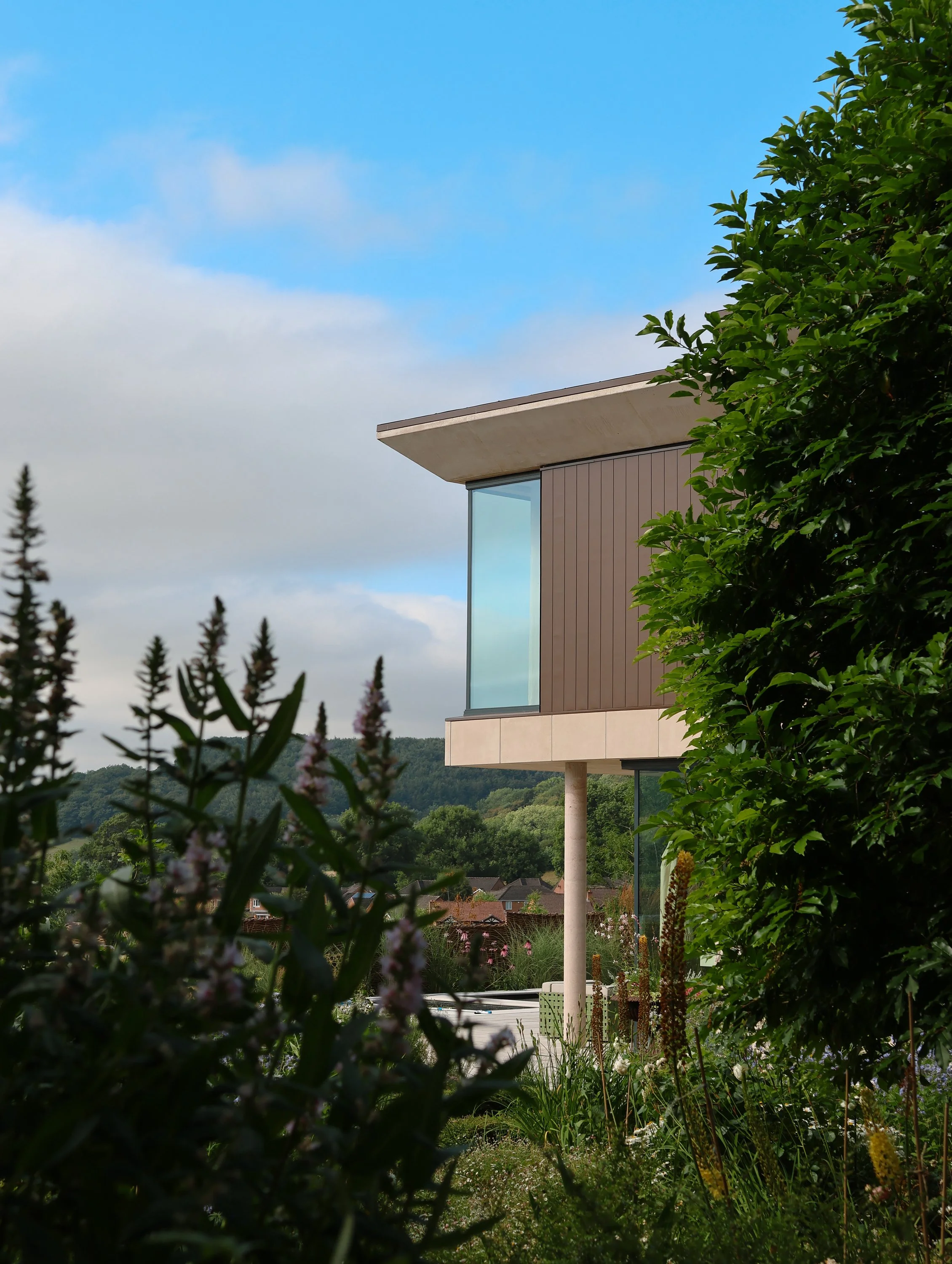 Modern house with large glass windows partially hidden behind lush green bushes, overlooking a village and hills under a partly cloudy sky.