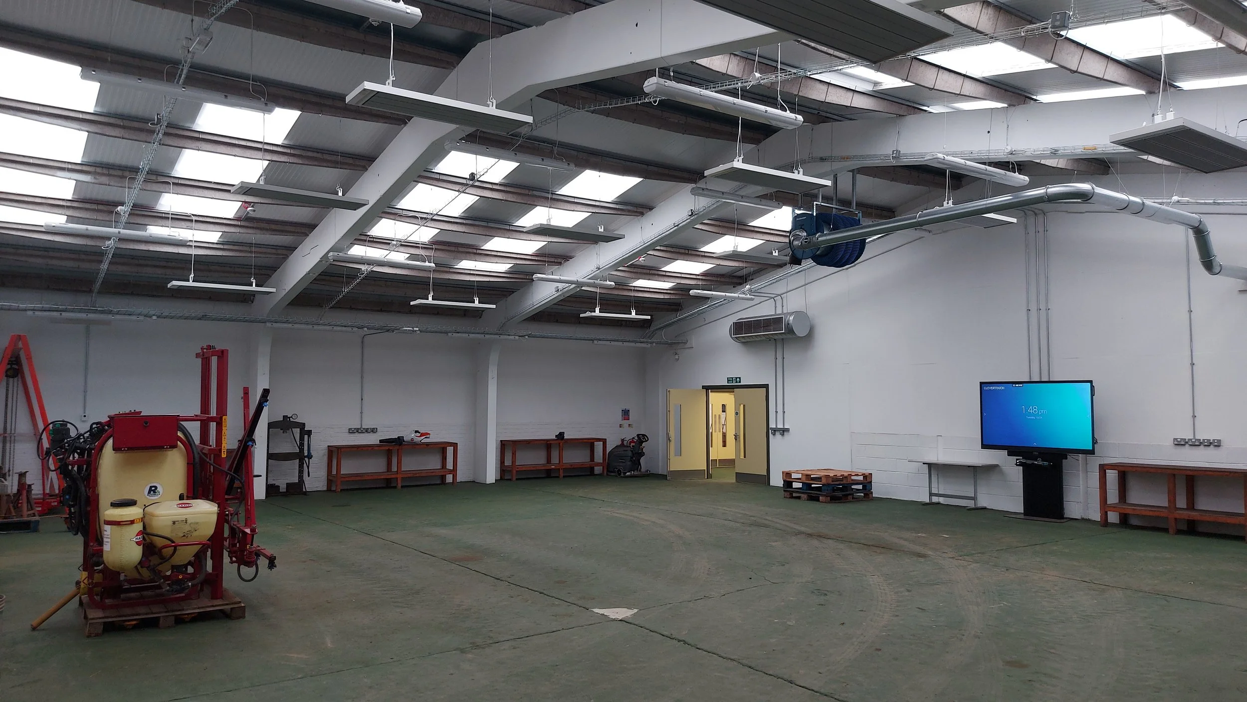 Empty industrial or workshop space with a green floor, white walls, and a high ceiling featuring skylights. There is machinery and equipment on the left, a TV monitor on a stand on the right, and a door in the back wall.