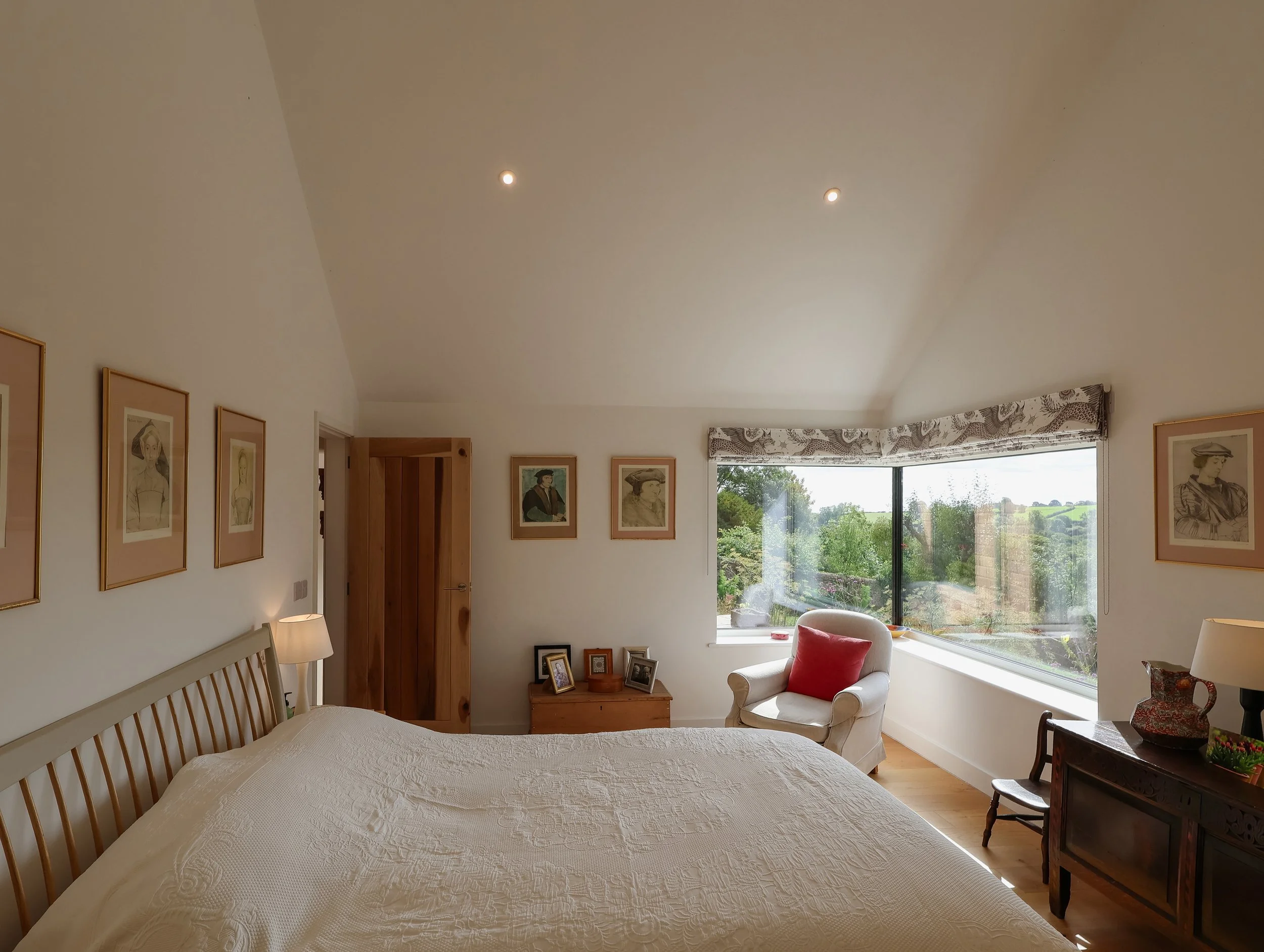 A cozy bedroom with a large bed, framed artwork on the walls, a window with a view of greenery, and a sitting area with an armchair and side table.