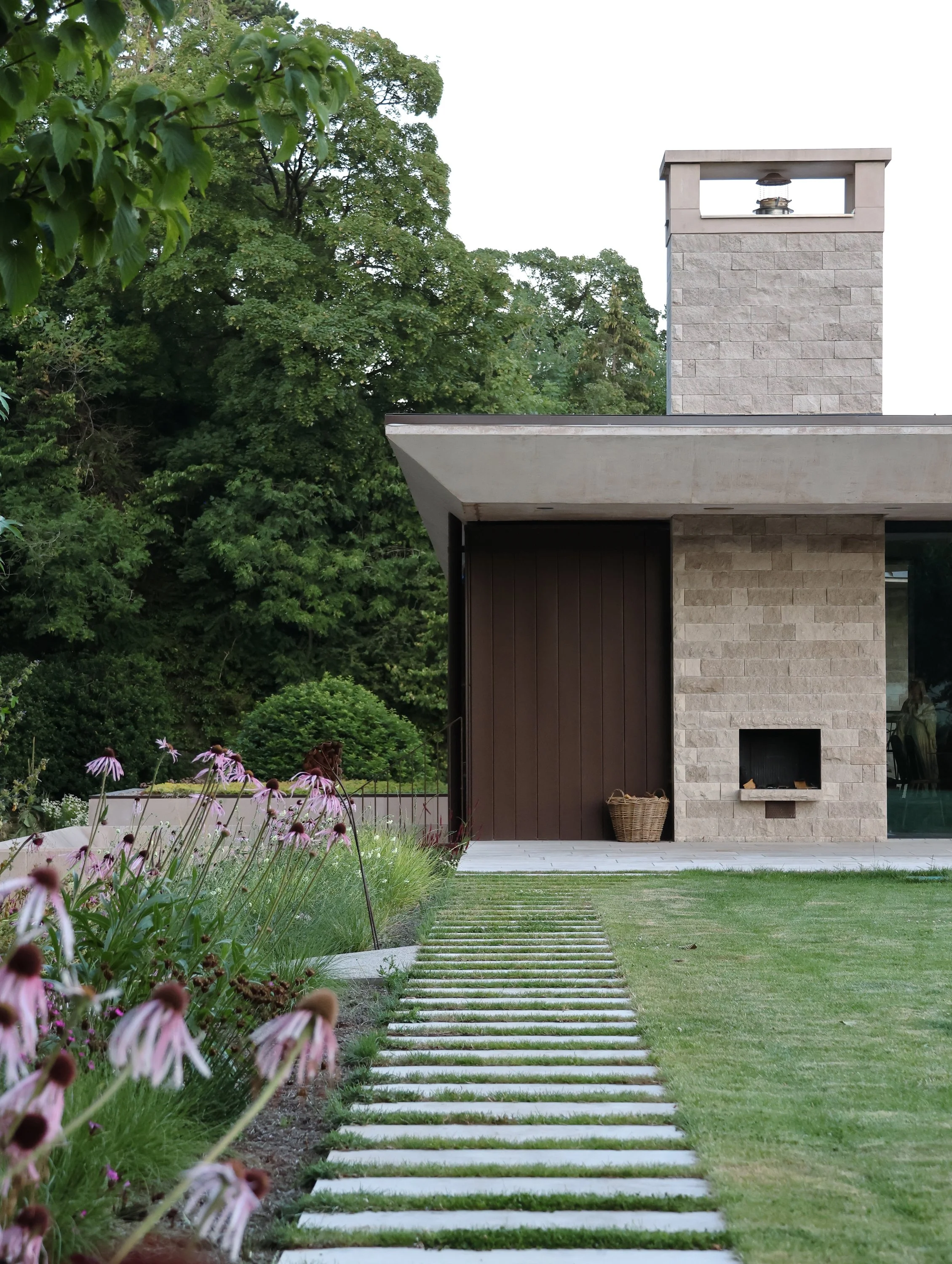 A modern home with a stone exterior and a chimney, surrounded by a well-maintained lawn and a garden with pink flowers, and a stone pathway leading to the house.