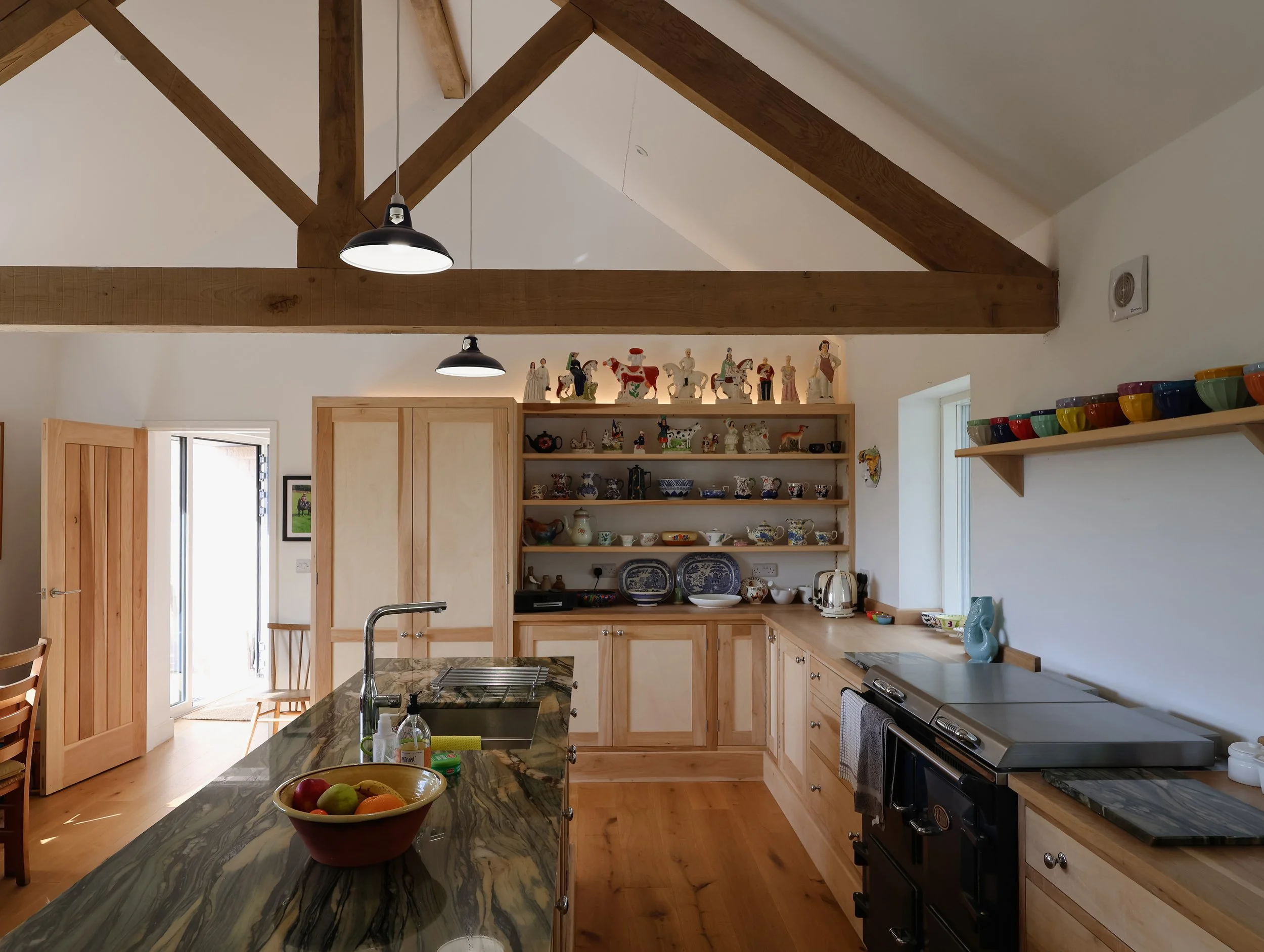 A kitchen with wooden cabinets, a marble countertop, and open shelves displaying decorative and collectible items. The ceiling has exposed wooden beams, with two black pendant lights hanging down. The space is bright with natural light coming in thro