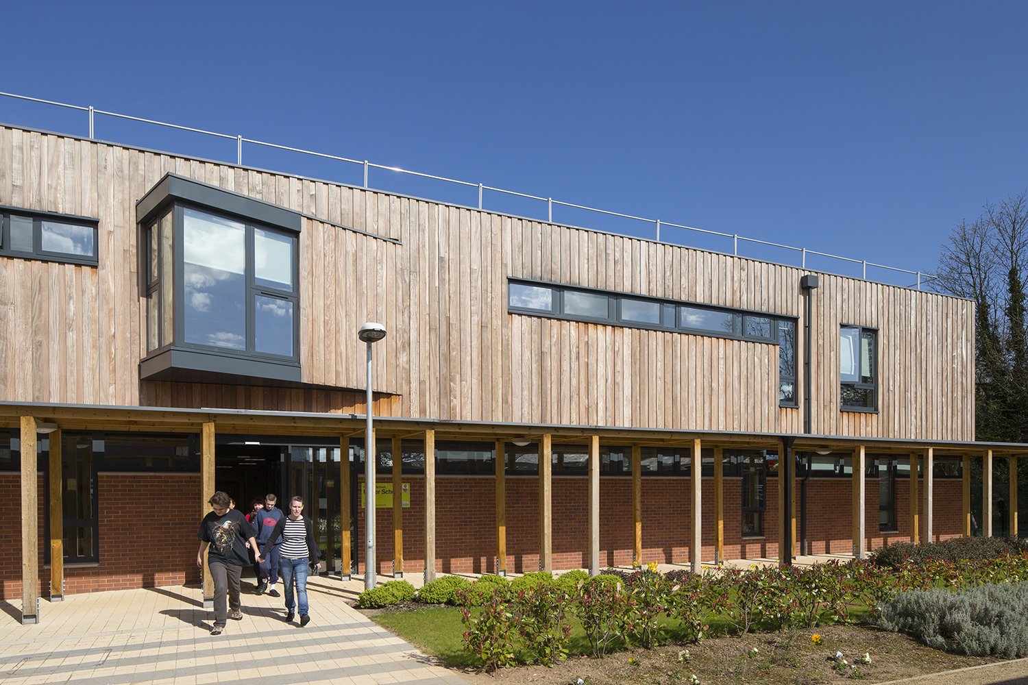 Students walking outside a modern school building with brick and wooden exterior and large windows, under a clear blue sky.