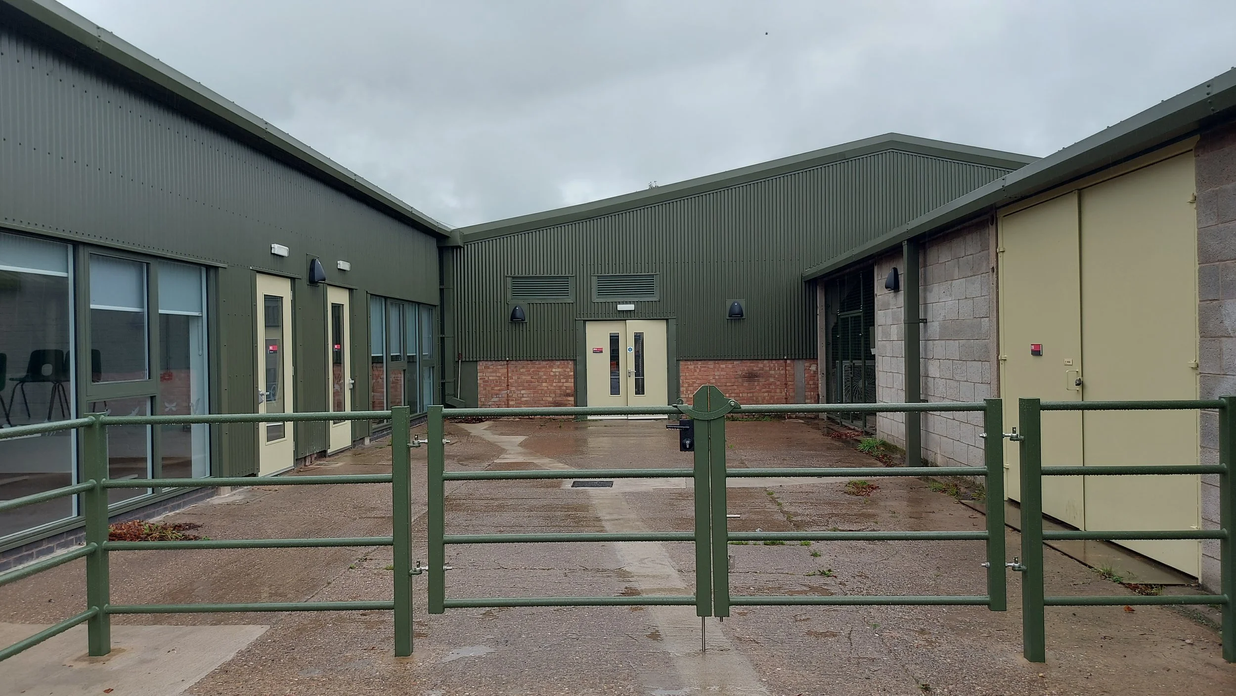 View of a modern commercial building with green metal siding, large windows, a white door, and outdoor lighting fixtures; surrounded by a green metal fence on a cloudy day.