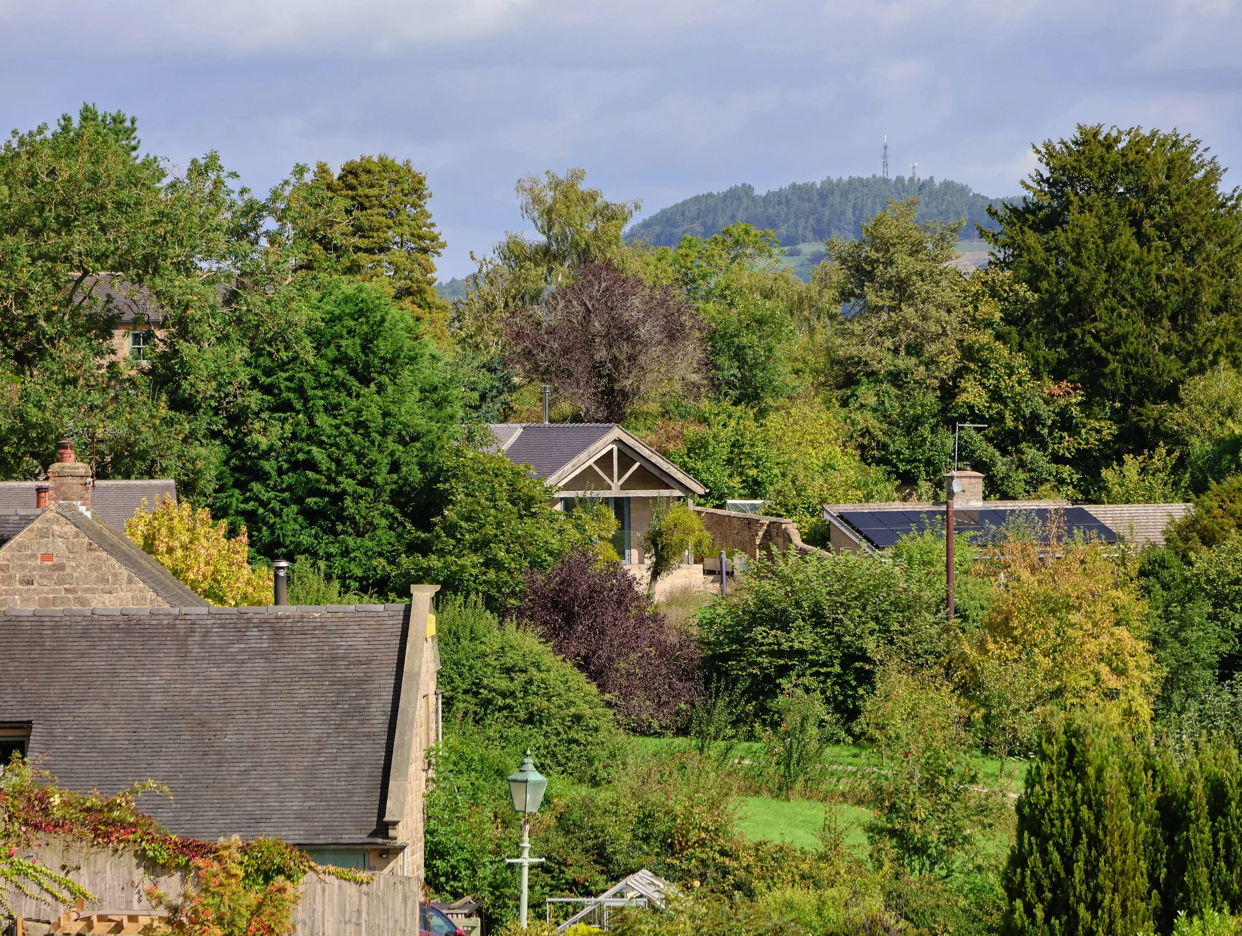 Residential area with rooftops and lush green trees, hills in the background, and a partly cloudy sky.