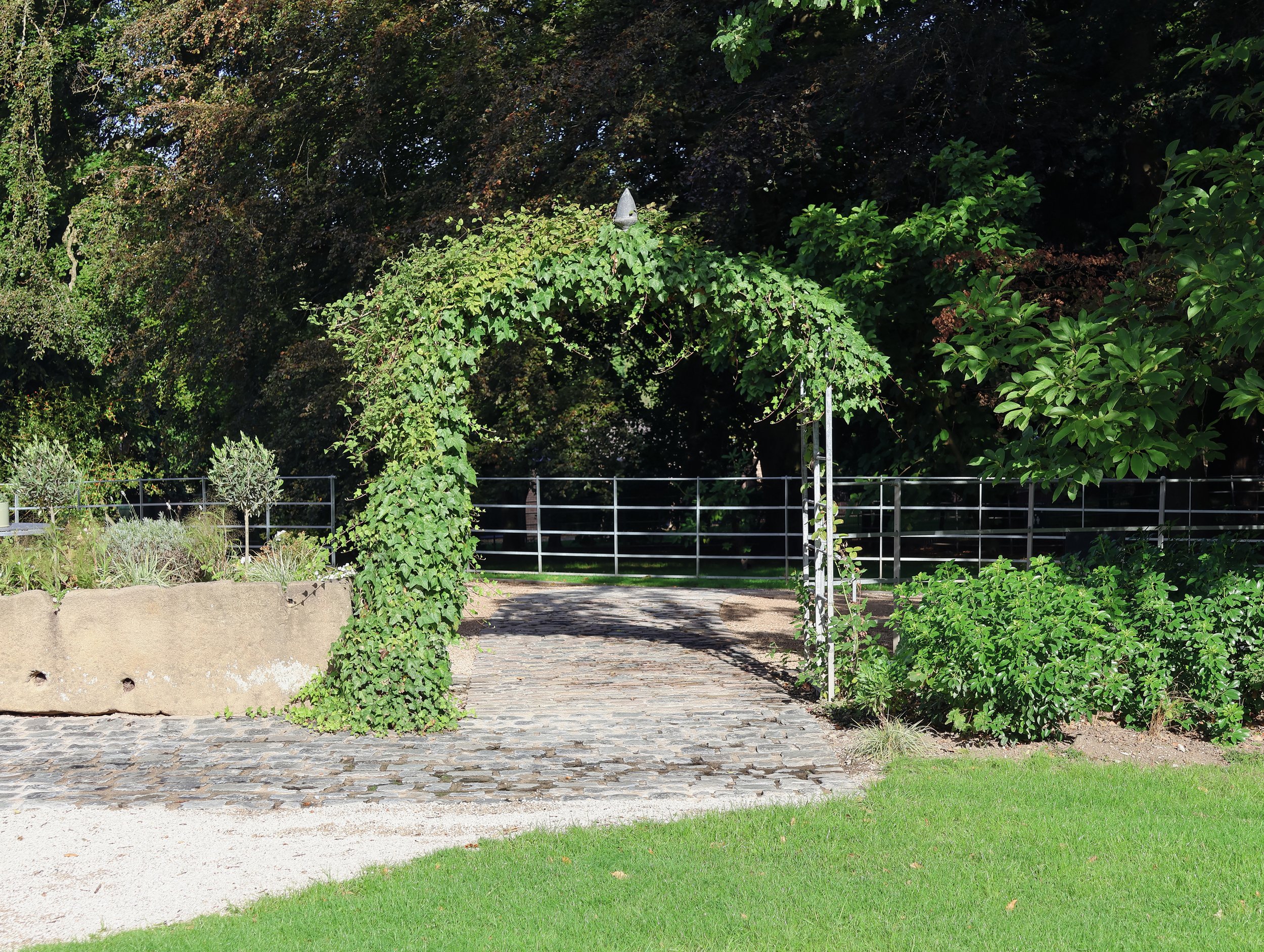 A park with a stone pathway, a metal fence and a variety of green trees and shrubs.