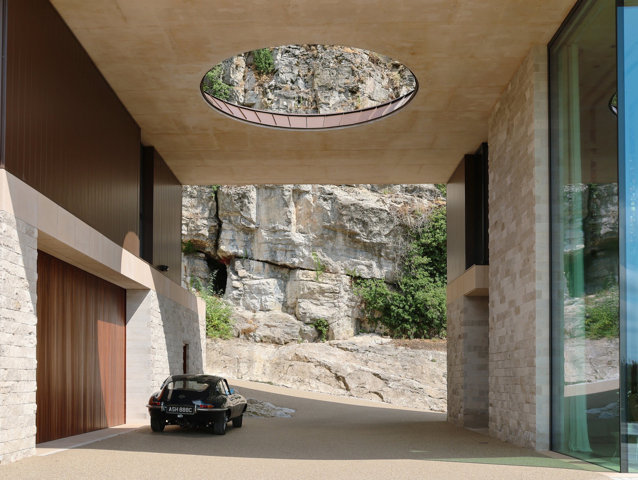 A modern building with stone and glass walls, a wooden garage door, and an oval skylight in the ceiling providing a view of rocky terrain and greenery above. A black vintage car is parked in the driveway.