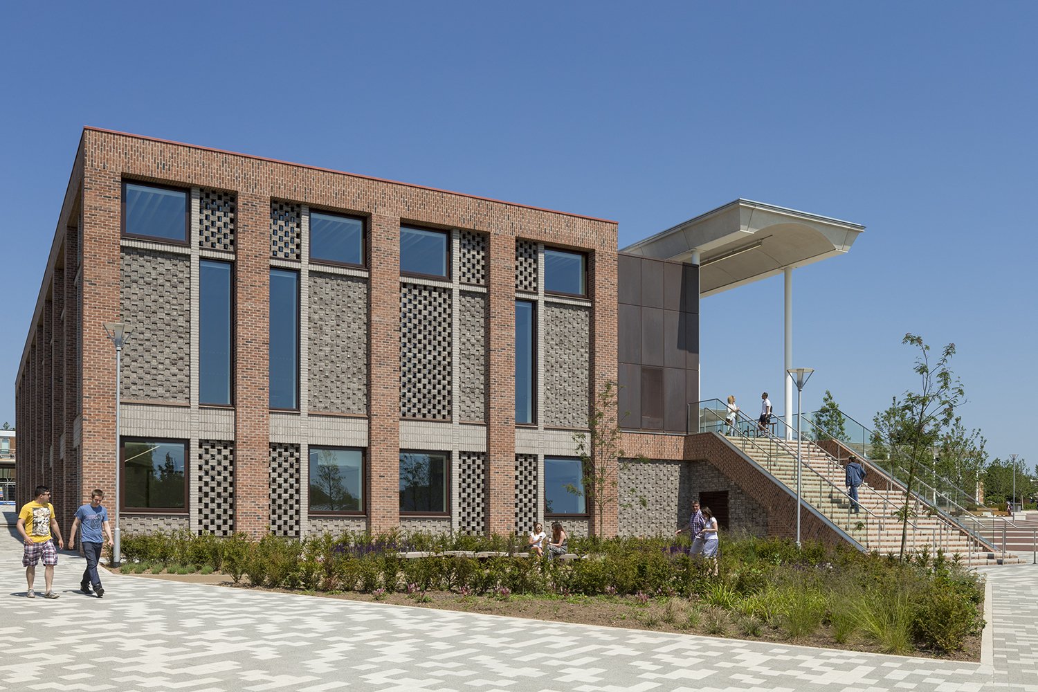 Modern brick and glass university building with outdoor staircase and people walking and sitting outside under a clear blue sky.