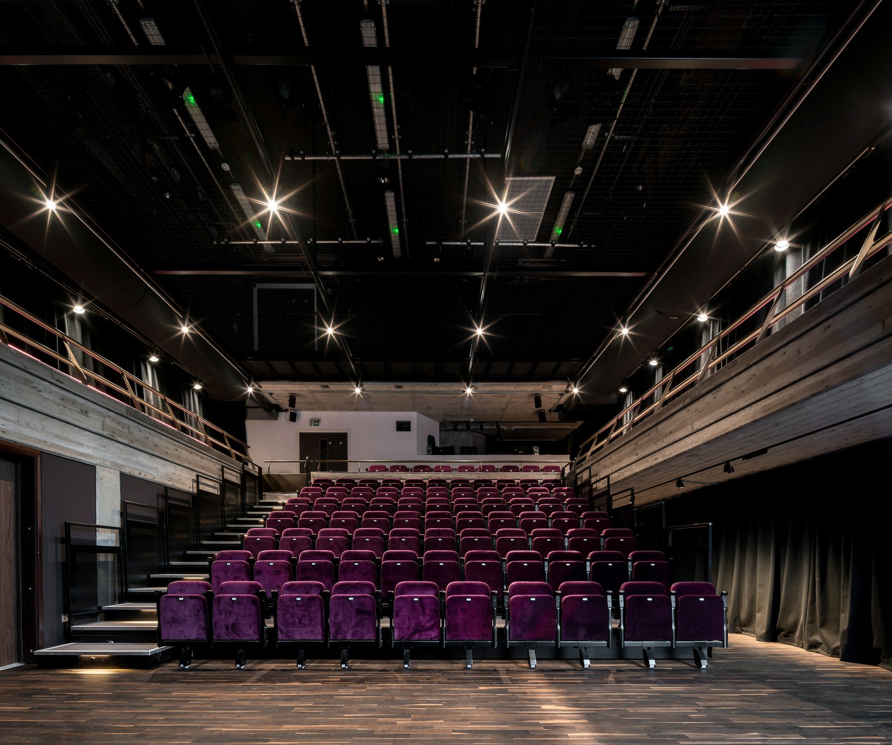 Empty theater with purple seats, wooden floors, black walls, a stage area, and black curtains on the sides, under a ceiling with bright lights and a grid structure.