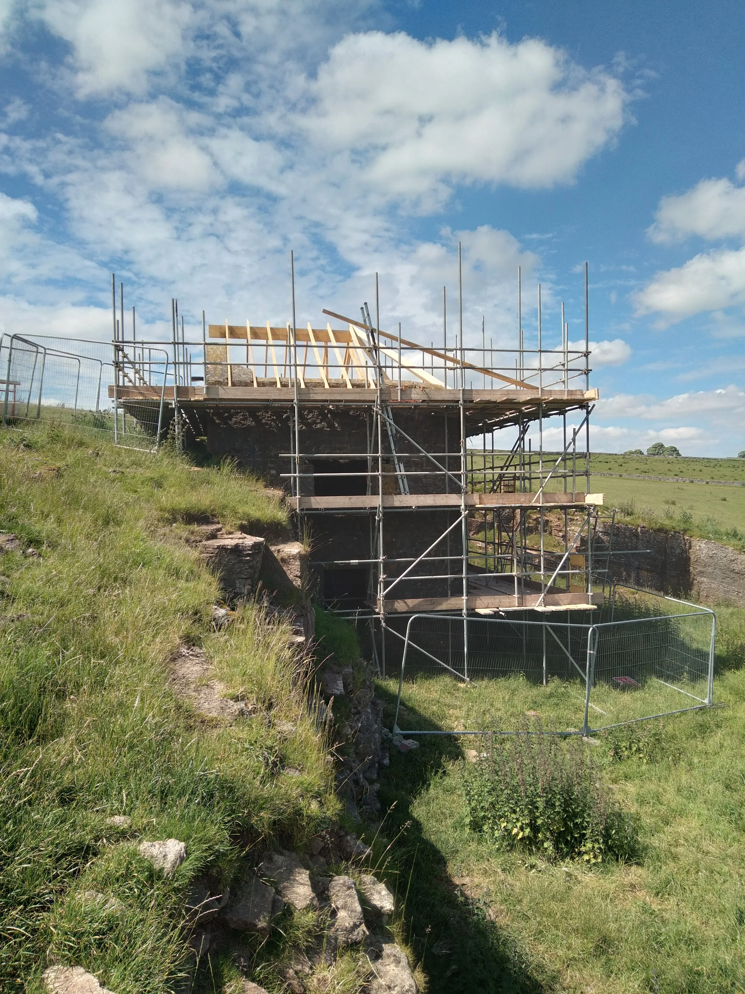 Under construction building with scaffolding on a grassy hillside, blue sky with scattered clouds in the background.