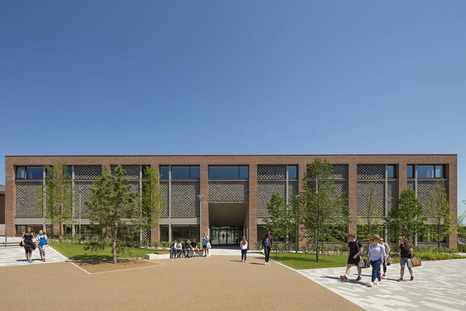 Students walking and sitting outside a modern university building with brick and concrete architecture, trees, and a clear blue sky.