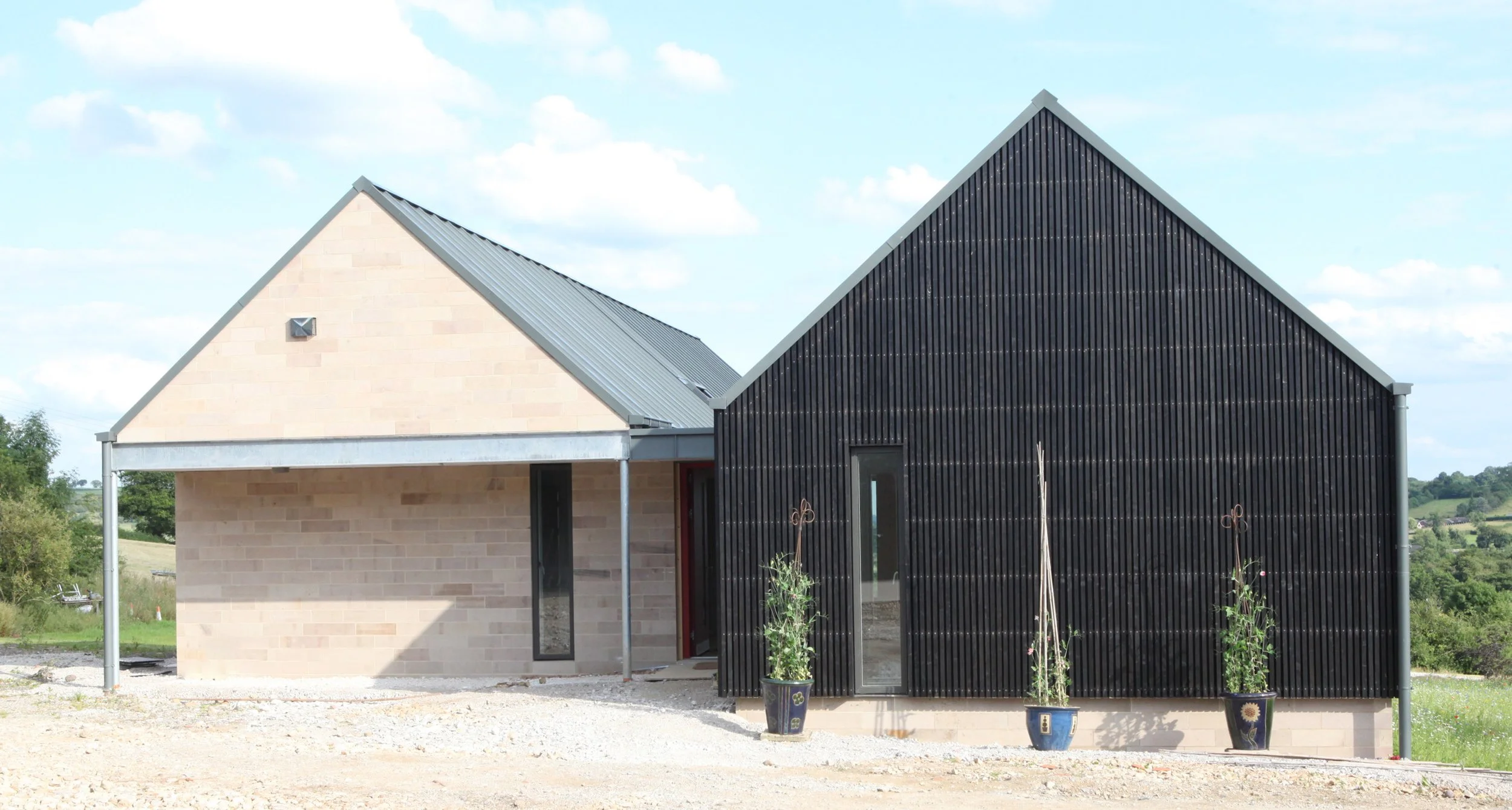 A modern house with a beige brick section and black wood-paneled section, gabled roof, in a rural setting with greenery and a partly cloudy sky.