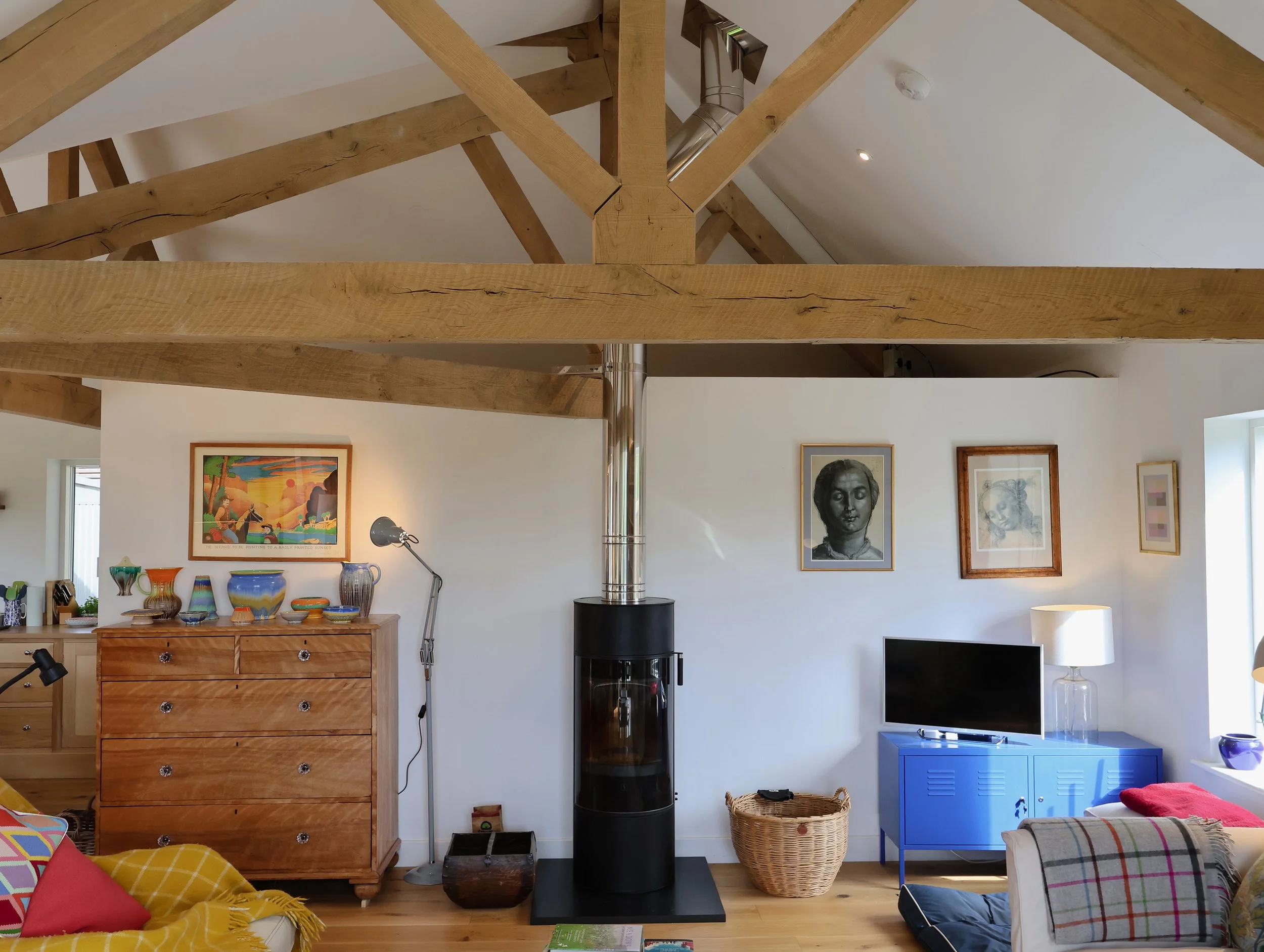 Interior of a living room with a high ceiling and wooden beams. A black stove is positioned against a white wall, with some artwork hanging above. There is a wooden chest of drawers on the left with glass vases, a table lamp, and decorative items. To