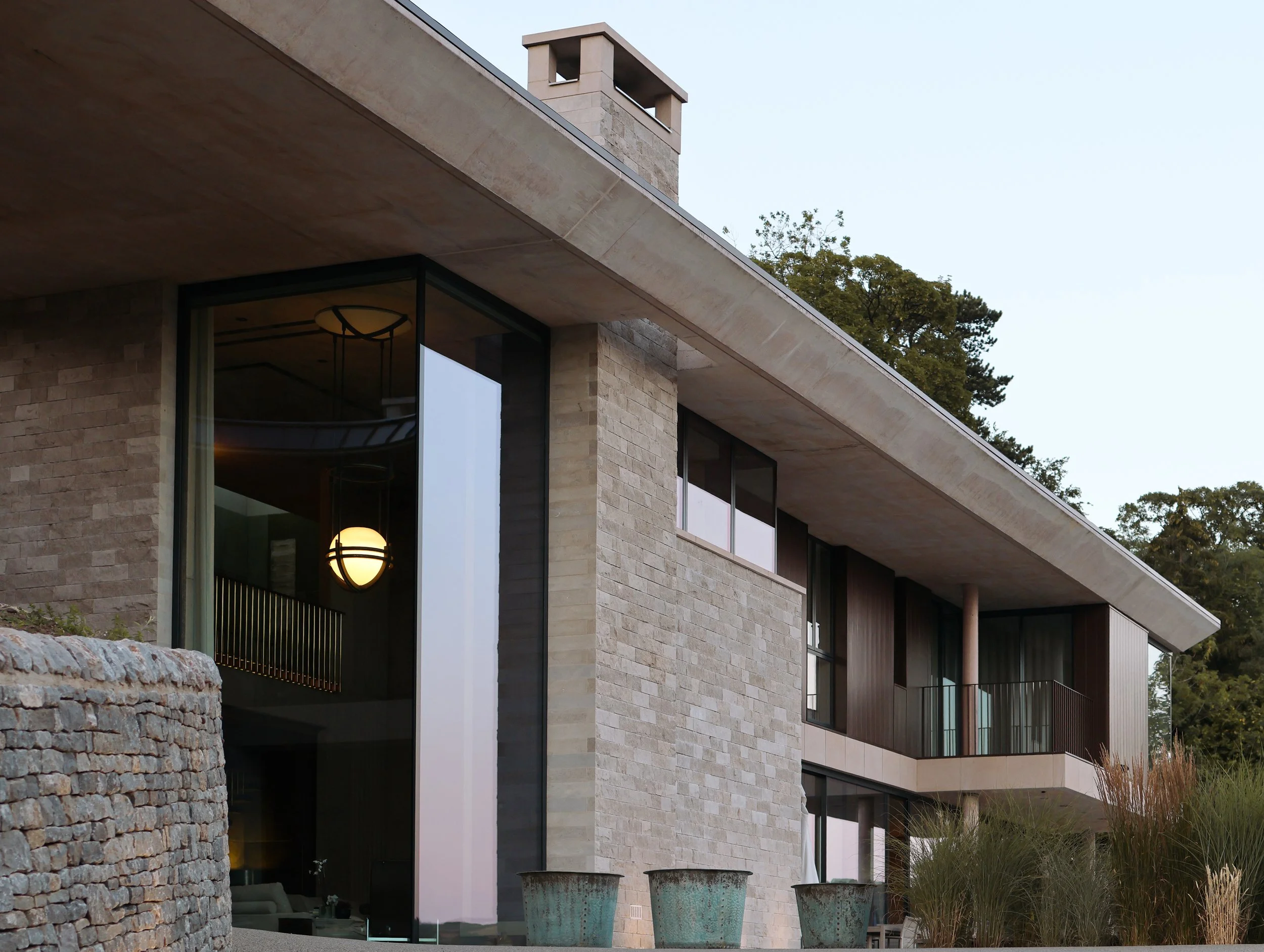 Contemporary multi-story house featuring large glass windows, a stone exterior, and a flat roof. The house is surrounded by trees and outdoor planters.