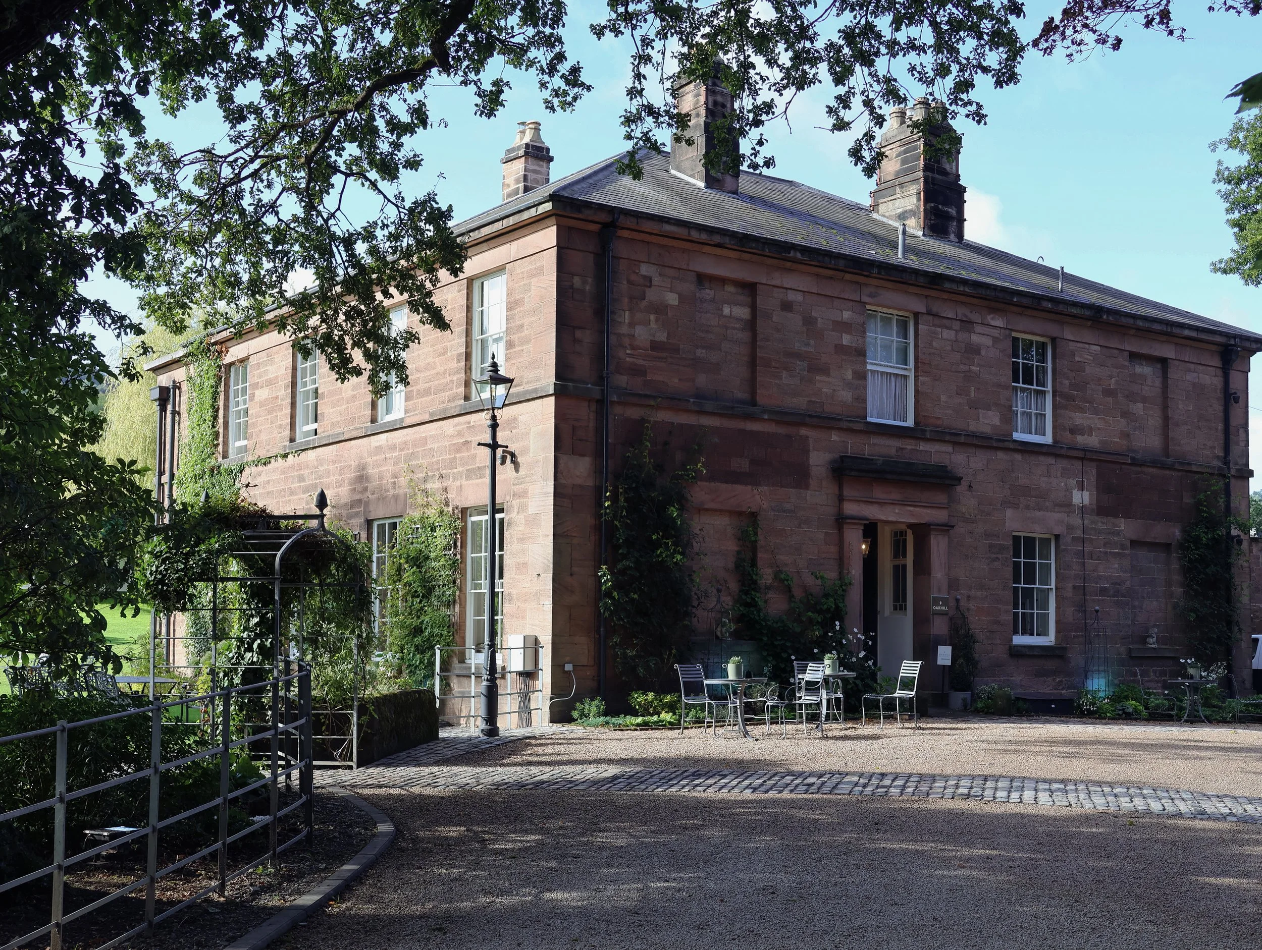 A large brick building with multiple chimneys and windows, surrounded by trees and a small patio with chairs, located in a park or garden setting.