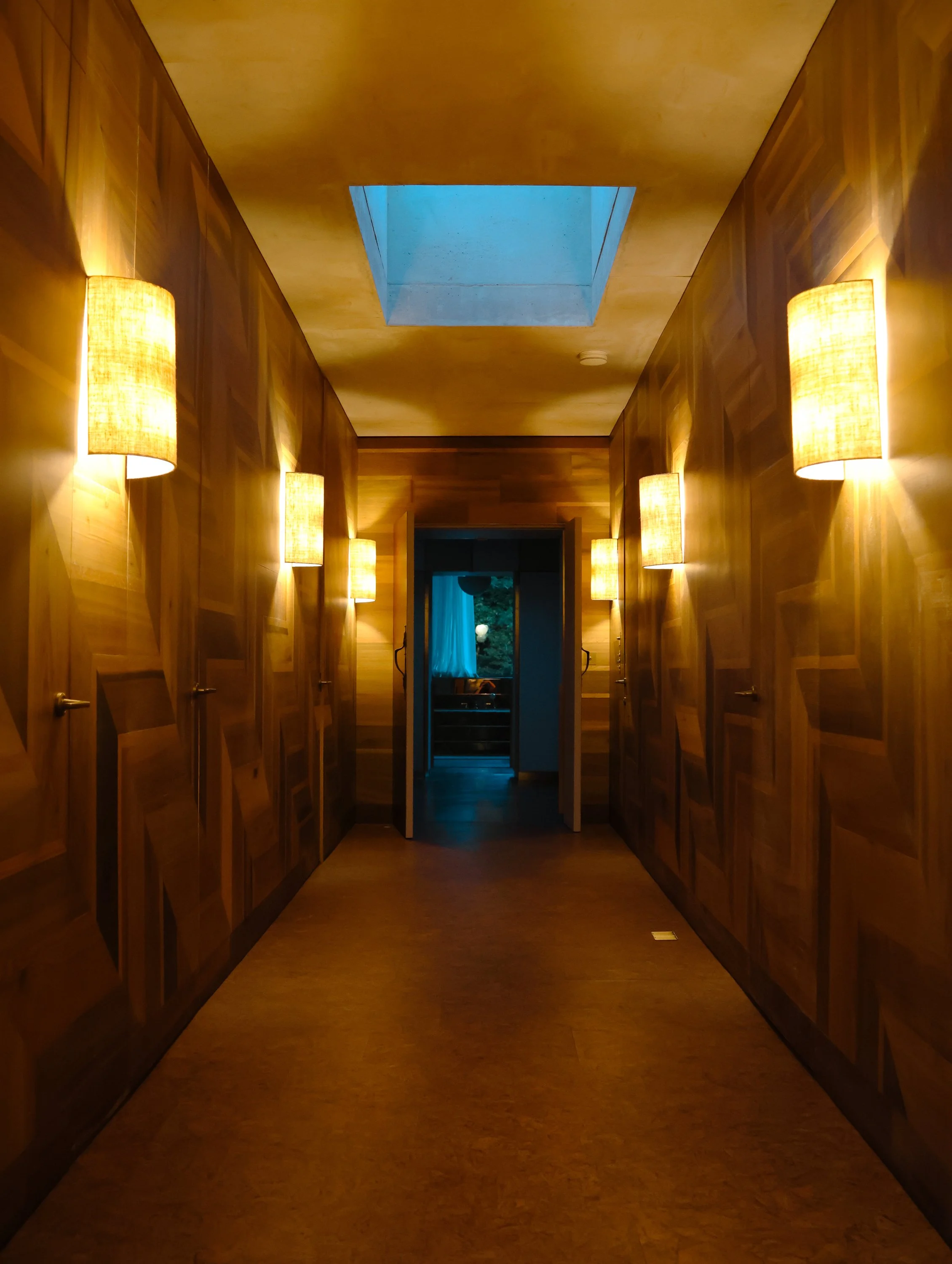 A hallway with wood-paneled walls and a ceiling, illuminated by wall-mounted lamps. A skylight in the ceiling allows natural light into the space. At the end of the hallway, there is an open door leading to a room with a window and visible outdoor gr