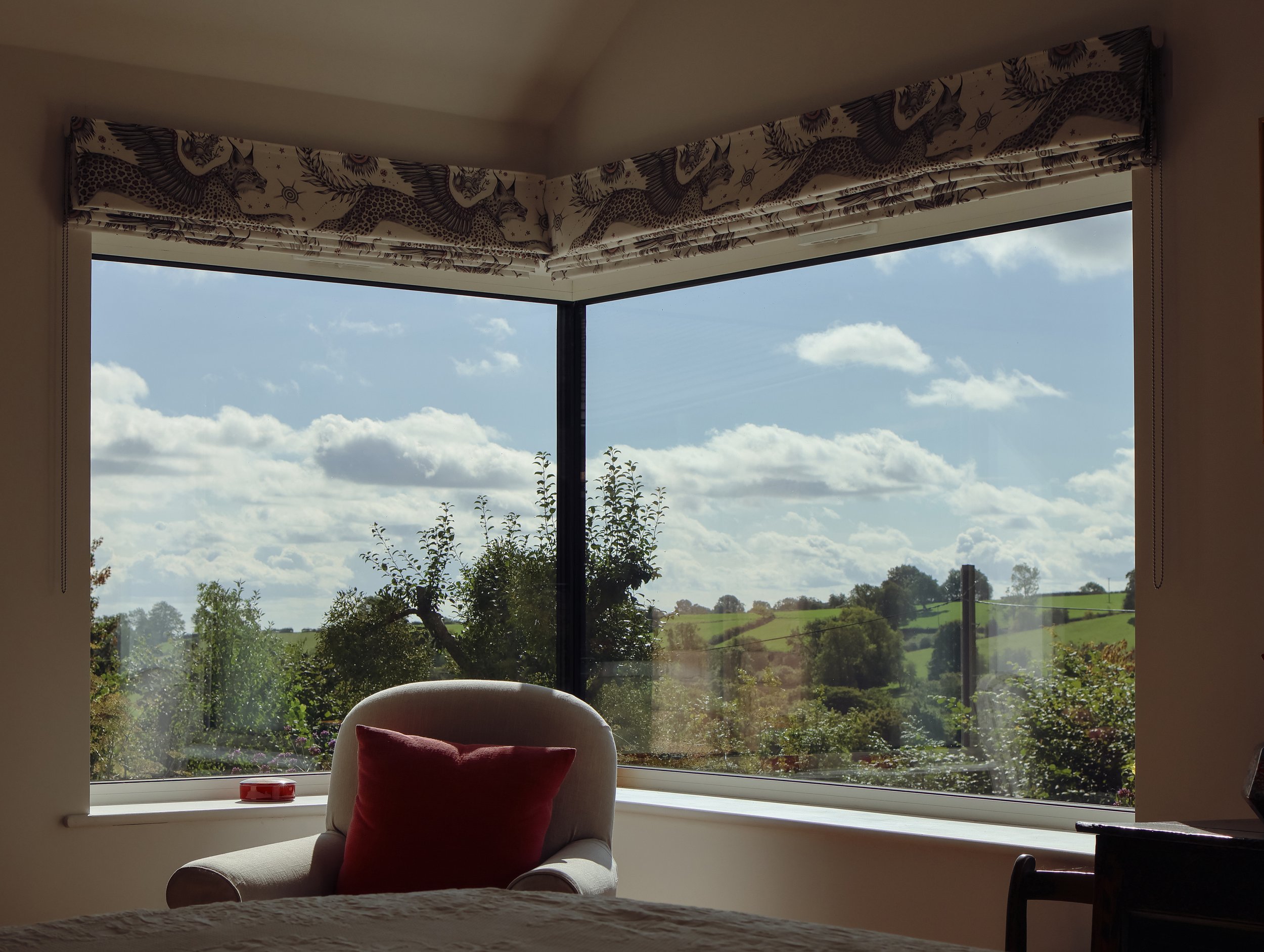 A cozy indoor space with a large window showing a scenic view of trees and a cloudy sky, with a white chair and a red pillow in the foreground.