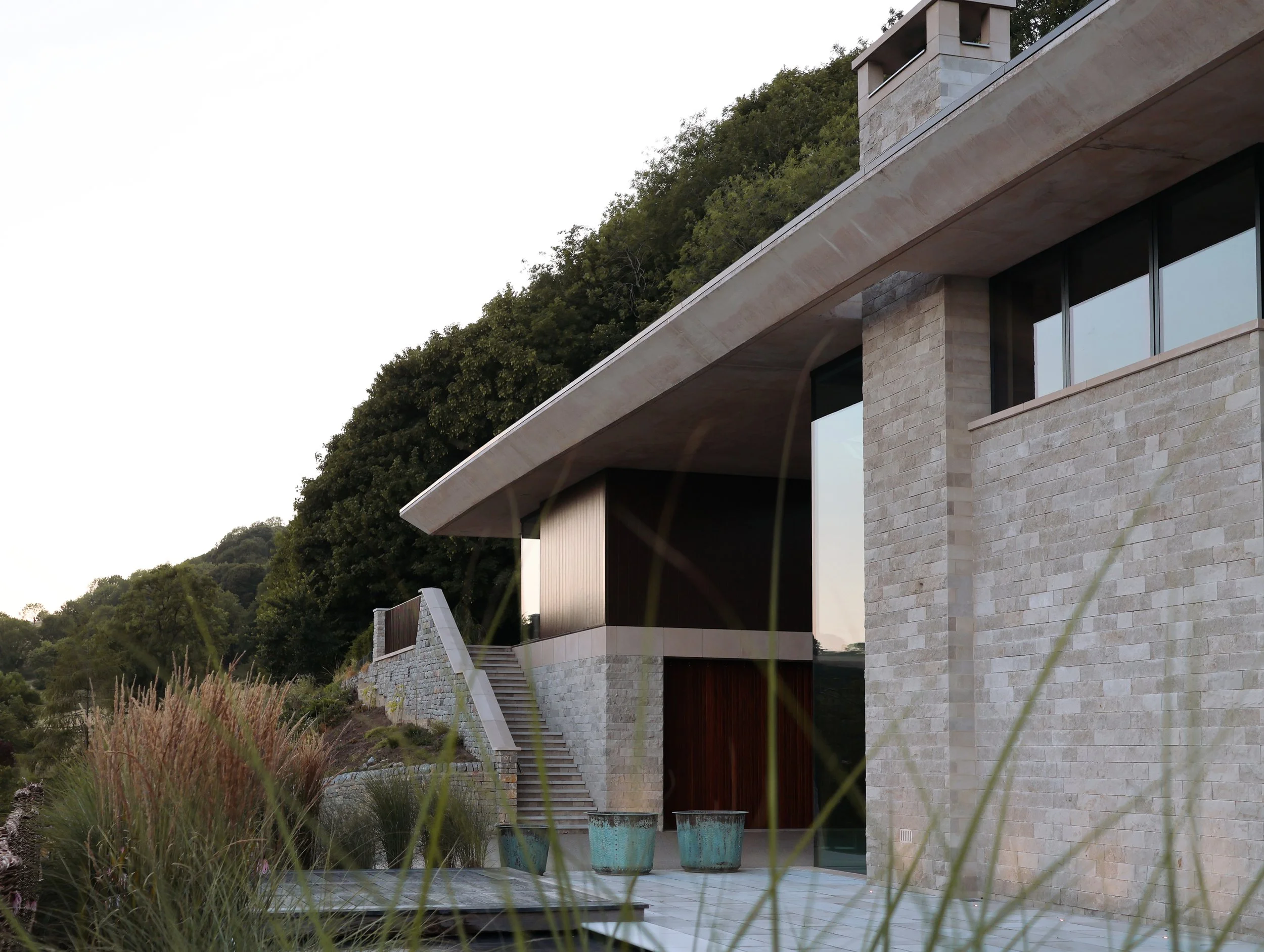 Modern house with stone exterior, large windows, and outdoor stairs leading to a balcony, surrounded by greenery.