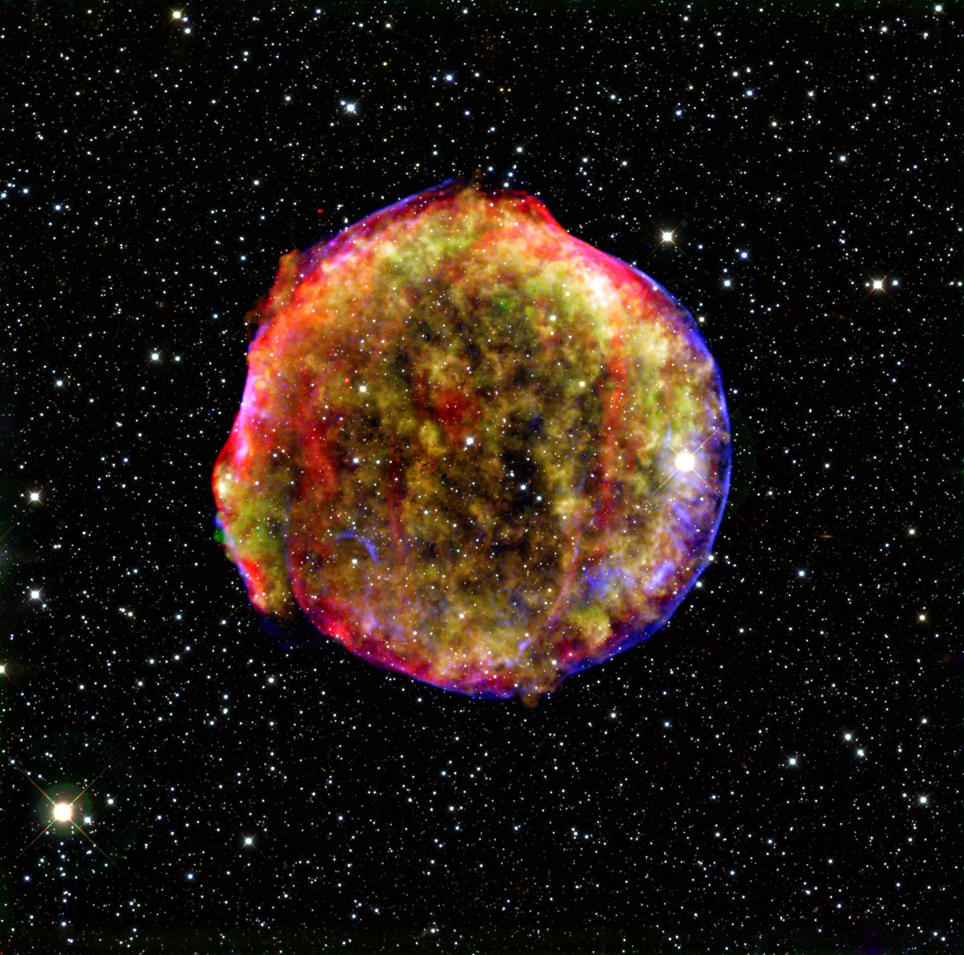 This composite image of a supernova remnant combines infrared and X-ray observations. The explosion left a blazing hot cloud of expanding debris (green and yellow). The location of the blast's outer shock wave can be seen as a blue sphere of ultra-energetic electrons. Newly synthesized dust in the ejected material and heated pre-existing dust from the area around the supernova radiate at infrared wavelengths of 24 microns (red). Foreground and background stars in the image are white. Image Credit: MPIA/NASA/Calar Alto Observatory.