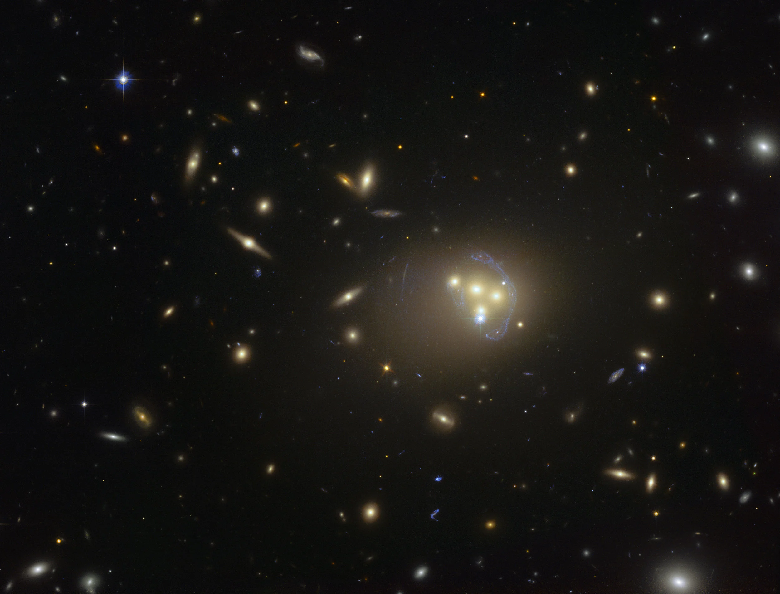 This image from the NASA/ESA Hubble Space Telescope shows the rich galaxy cluster Abell 3827. The strange blue structures surrounding the central galaxies are gravitationally lensed views of a much more distant galaxy behind the cluster. Observations of the central four merging galaxies have provided hints that the dark matter around one of the galaxies is not moving with the galaxy itself, possibly implying dark matter-dark matter interactions of an unknown nature are occuring. Image credit: ESO