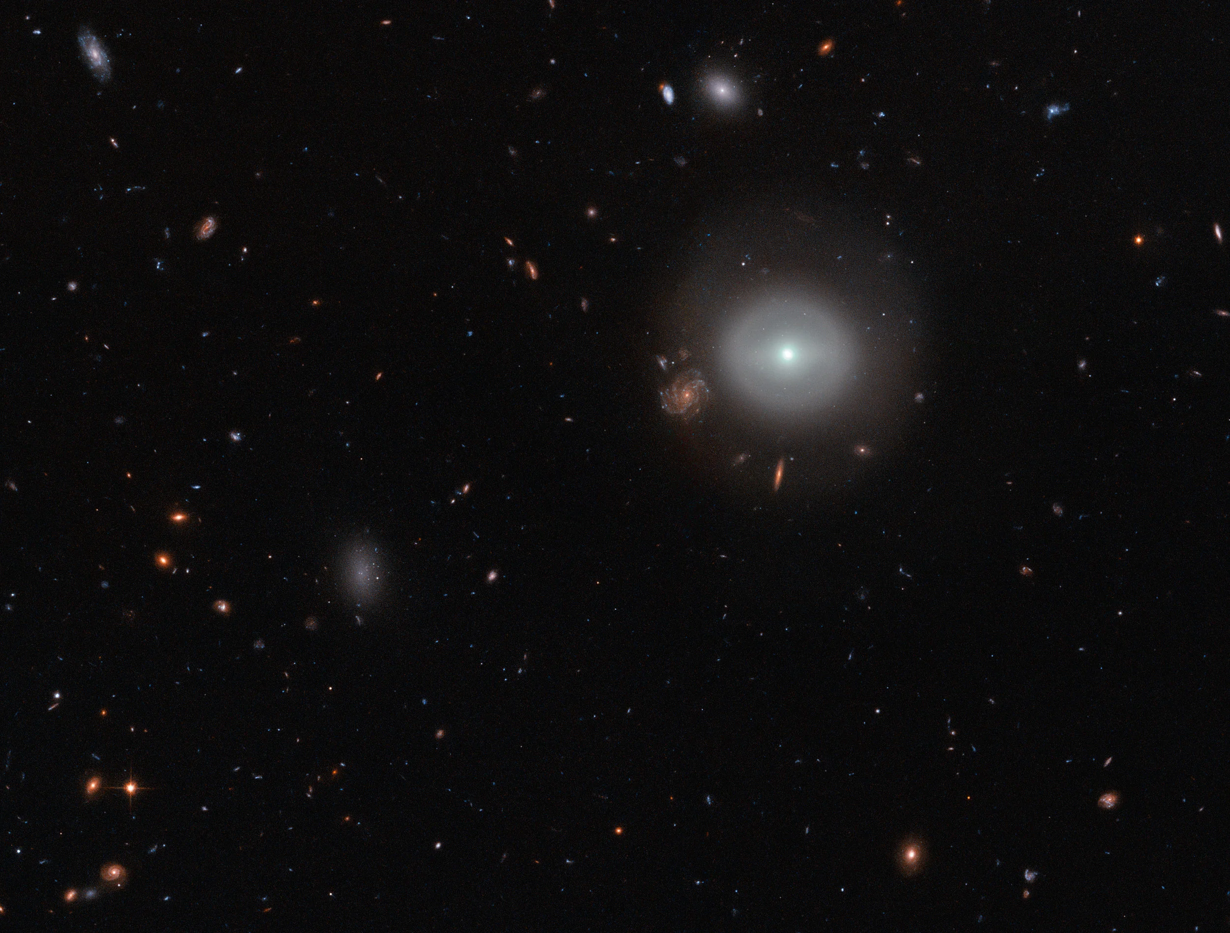 A lone source shines out brightly from the dark expanse of deep space, glowing softly against a picturesque backdrop of distant stars and colorful galaxies. This scene shows PGC 83677, a lenticular galaxy — a galaxy type that sits between the more familiar elliptical and spiral varieties in the Hubble sequence. Image credit: ESA/Hubble & NASA | Acknowledgements: Judy Schmidt (Geckzilla)